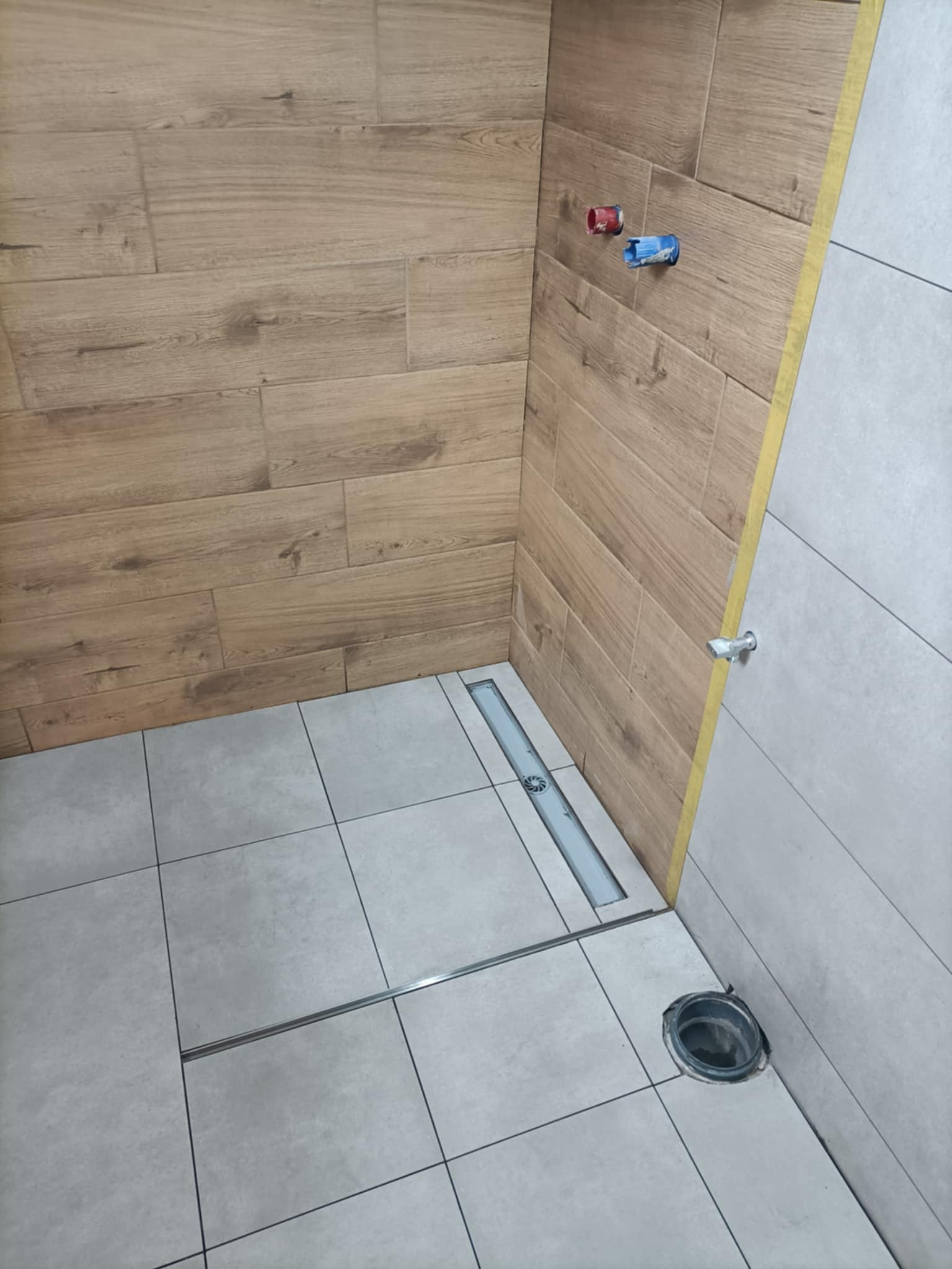 Nowoczesna łazienka z drewnopodobnymi płytkami ściennymi i szarymi podłogowymi. Widoczny odpływ liniowy i przygotowane przyłącza wodne.