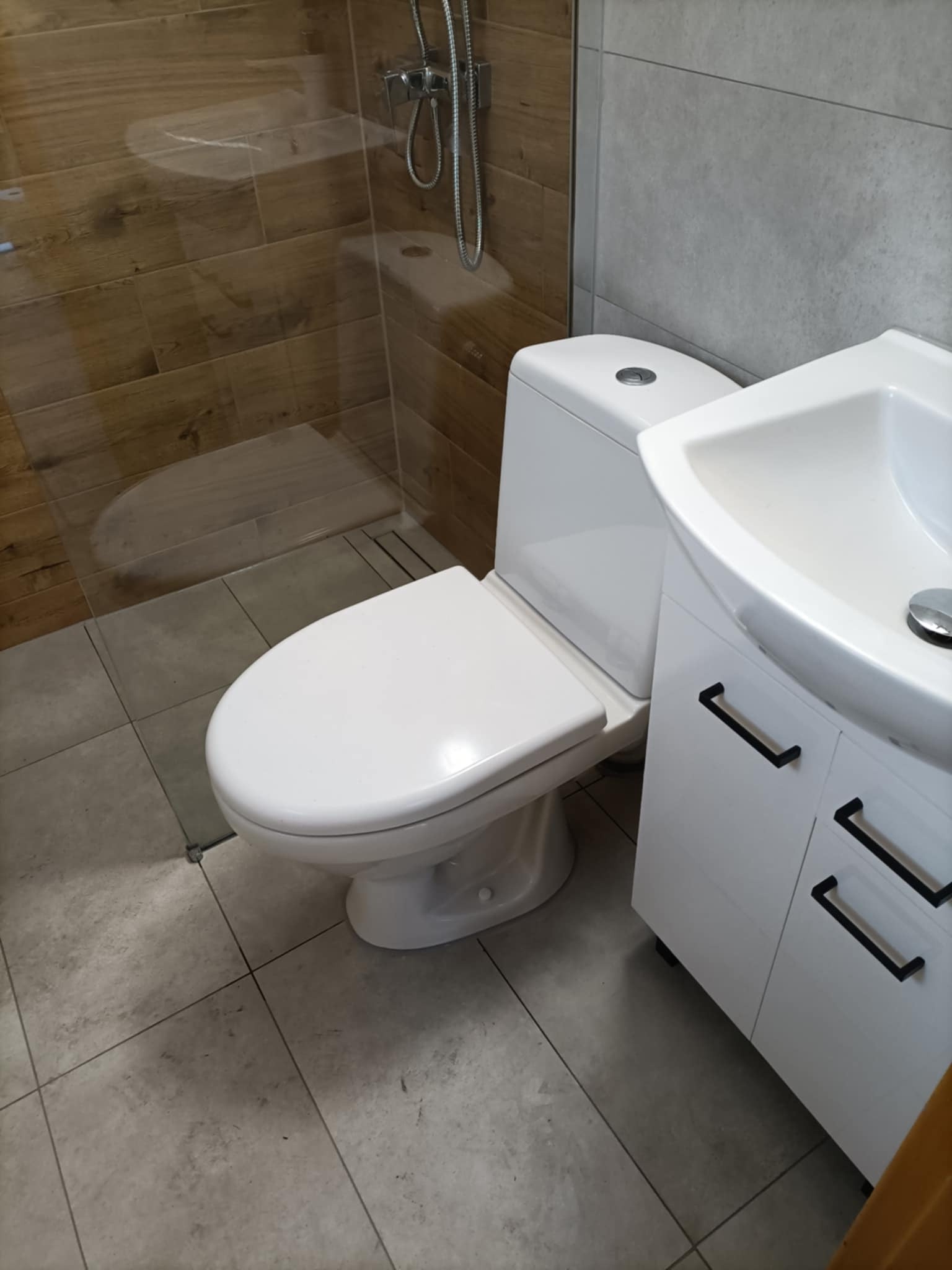 Biała toaleta i umywalka z szafką w wykończonej łazience. Widoczne płytki imitujące drewno pod prysznicem i szare płytki na podłodze.