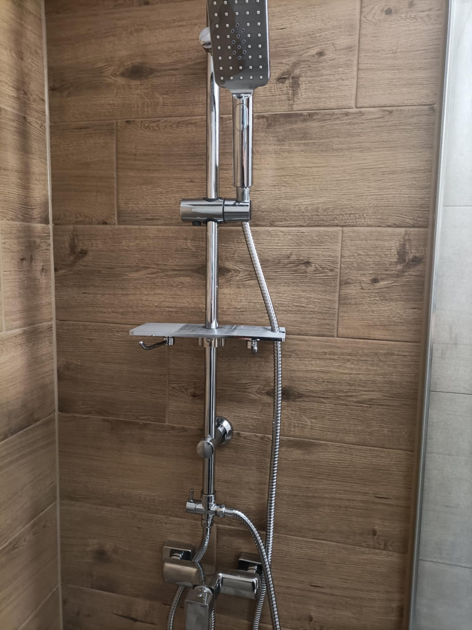 Chromowany zestaw natryskowy z deszczownicą na tle ściany z płytek imitujących drewno. Widoczny fragment kabiny prysznicowej z szarych płytek.