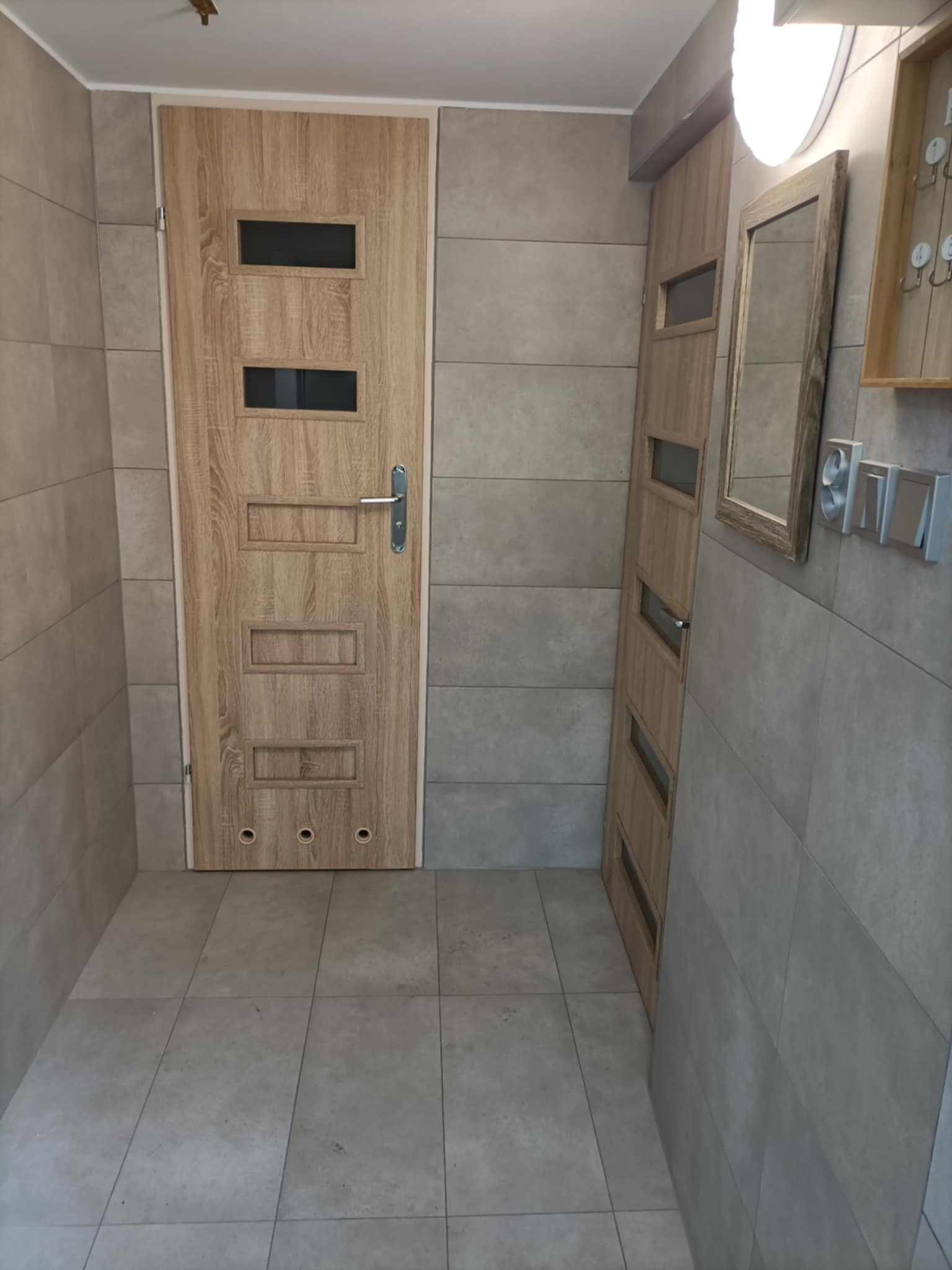 Wykończone wnętrze przedpokoju: szare płytki na ścianach i podłodze, drewnopodobne drzwi z przeszkleniami, lustro w drewnianej ramie i włączniki światła.