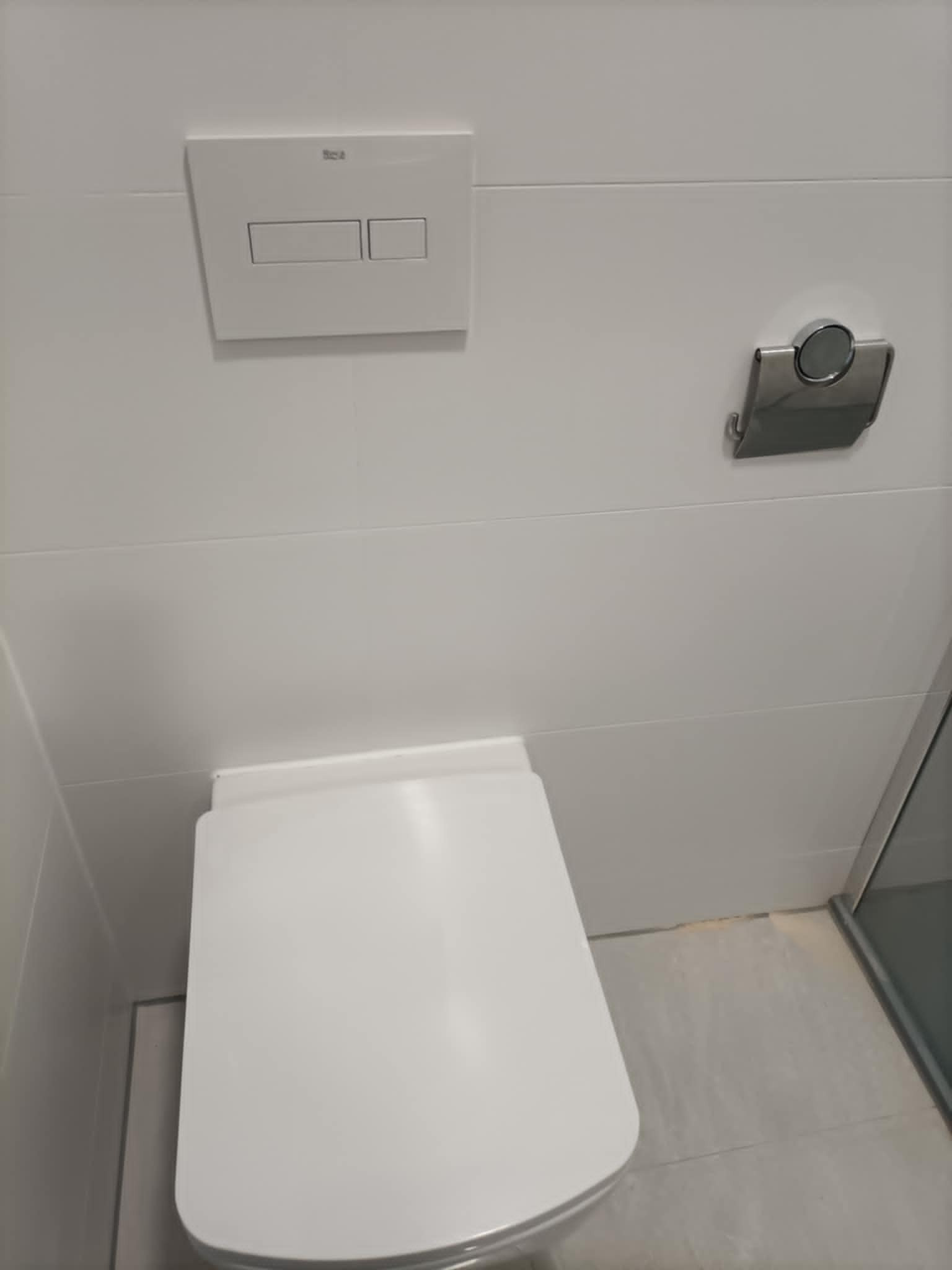 Biała toaleta podwieszana z dwudzielnym przyciskiem spłukującym Roca na tle białych płytek w minimalistycznej łazience. Widoczny uchwyt na papier toaletowy.