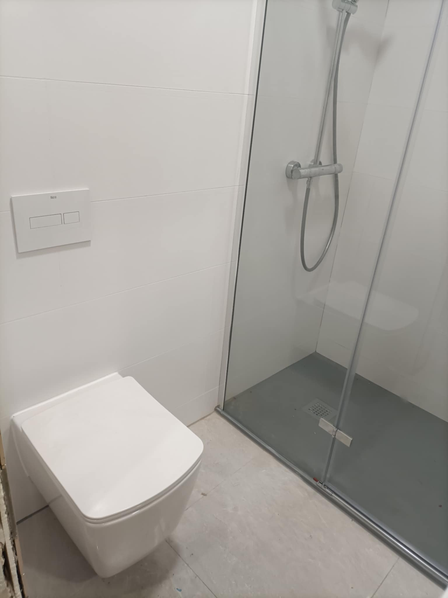 Nowoczesna, minimalistyczna łazienka z podwieszaną toaletą Roca i kabiną prysznicową ze szklaną ścianą. Białe płytki na ścianach i szary brodzik.