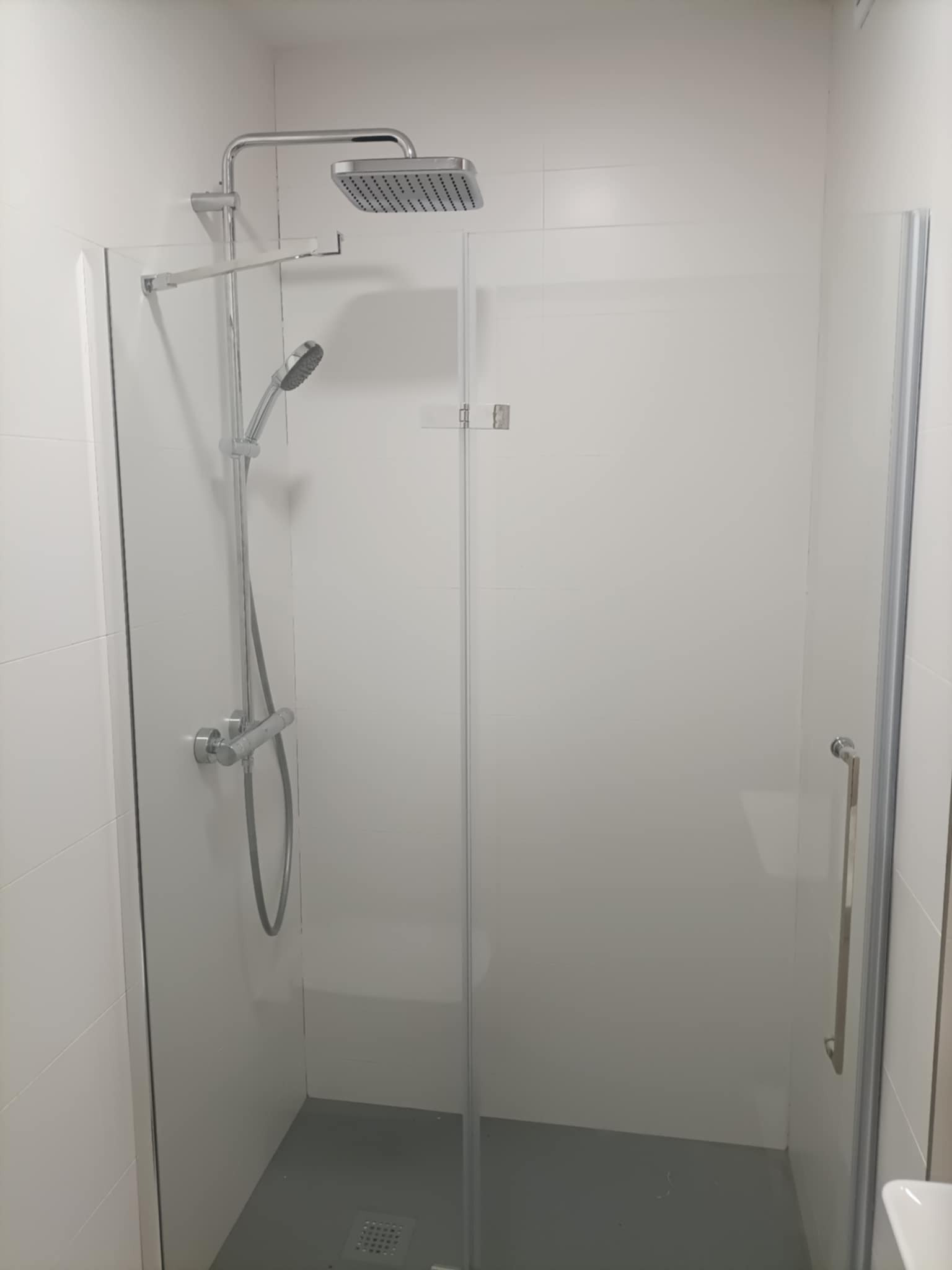 Nowoczesna kabina prysznicowa z deszczownicą i szklanymi drzwiami, wykończona białymi płytkami. Widoczny odpływ liniowy w szarej posadzce.