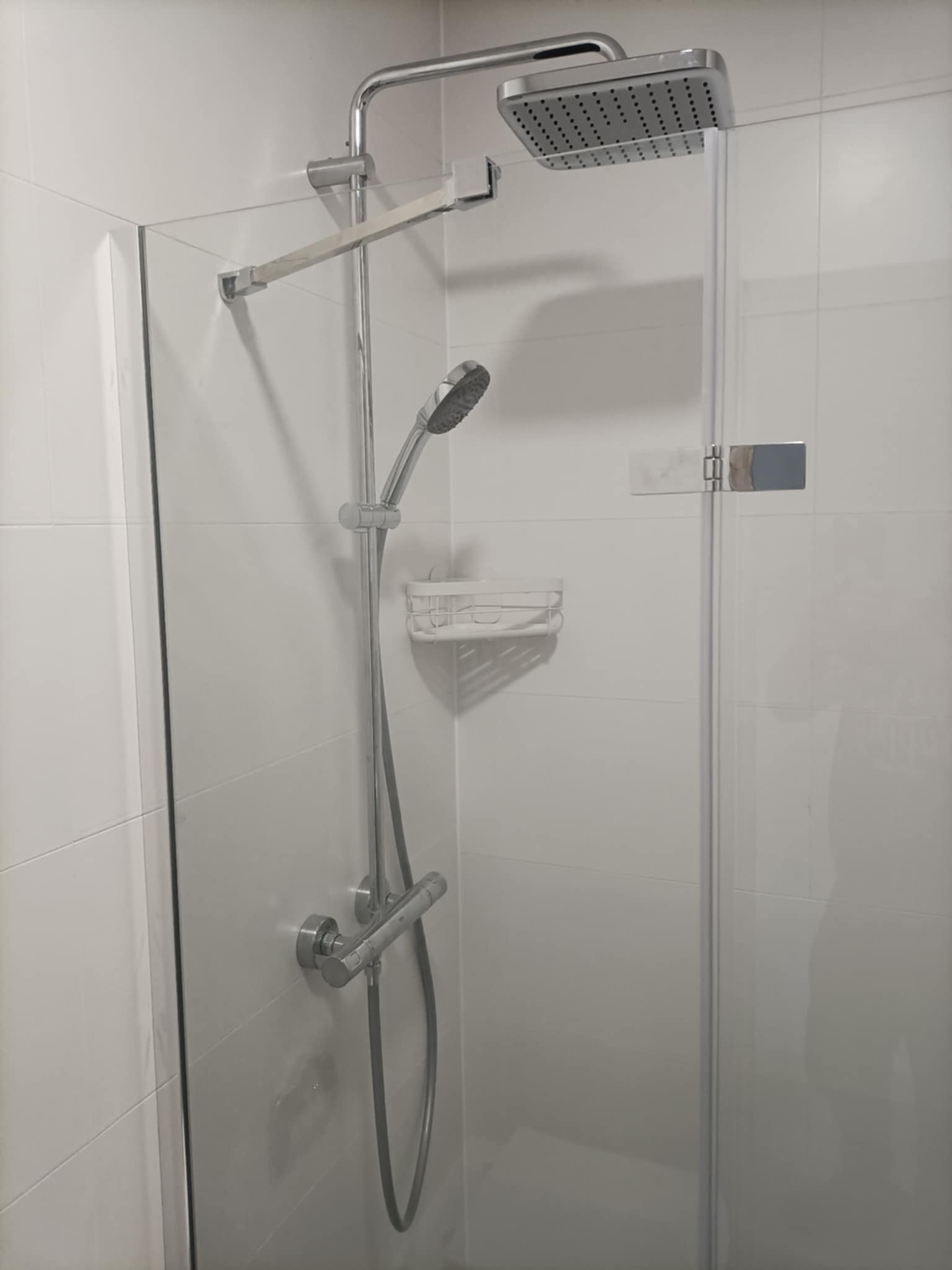 Nowoczesna kabina prysznicowa z deszczownicą i słuchawką, wykończona białymi płytkami. Minimalistyczny design, chromowane elementy, szklane drzwi z zawiasem.