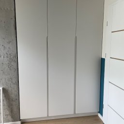 CAMELO Sandra Matyszkiewicz - Trzydrzwiowa szafa wnękowa z białymi frontami, widoczna surowa ściana betonowa po lewej stronie i fragment drzwi po prawej stronie na tle drewnianej podłogi.