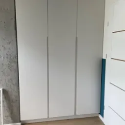 Trzydrzwiowa szafa wnękowa z białymi frontami, widoczna surowa ściana betonowa po lewej stronie i fragment drzwi po prawej stronie na tle drewnianej podłogi.