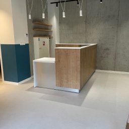 CAMELO Sandra Matyszkiewicz - Recepcja z blatem w kolorze białym i frontem w okleinie drewnopodobnej, widoczna na tle surowej, betonowej ściany i wiszących lamp