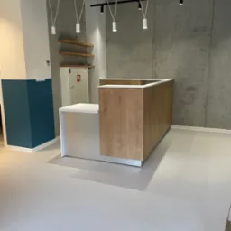 Recepcja z blatem w kolorze białym i frontem w okleinie drewnopodobnej, widoczna na tle surowej, betonowej ściany i wiszących lamp