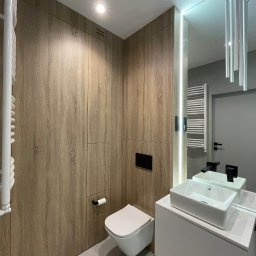 CAMELO Sandra Matyszkiewicz - Nowoczesna łazienka z drewnianą zabudową meblową, podwieszaną toaletą i białą umywalką na szafce, oświetlenie LED przy lustrze.