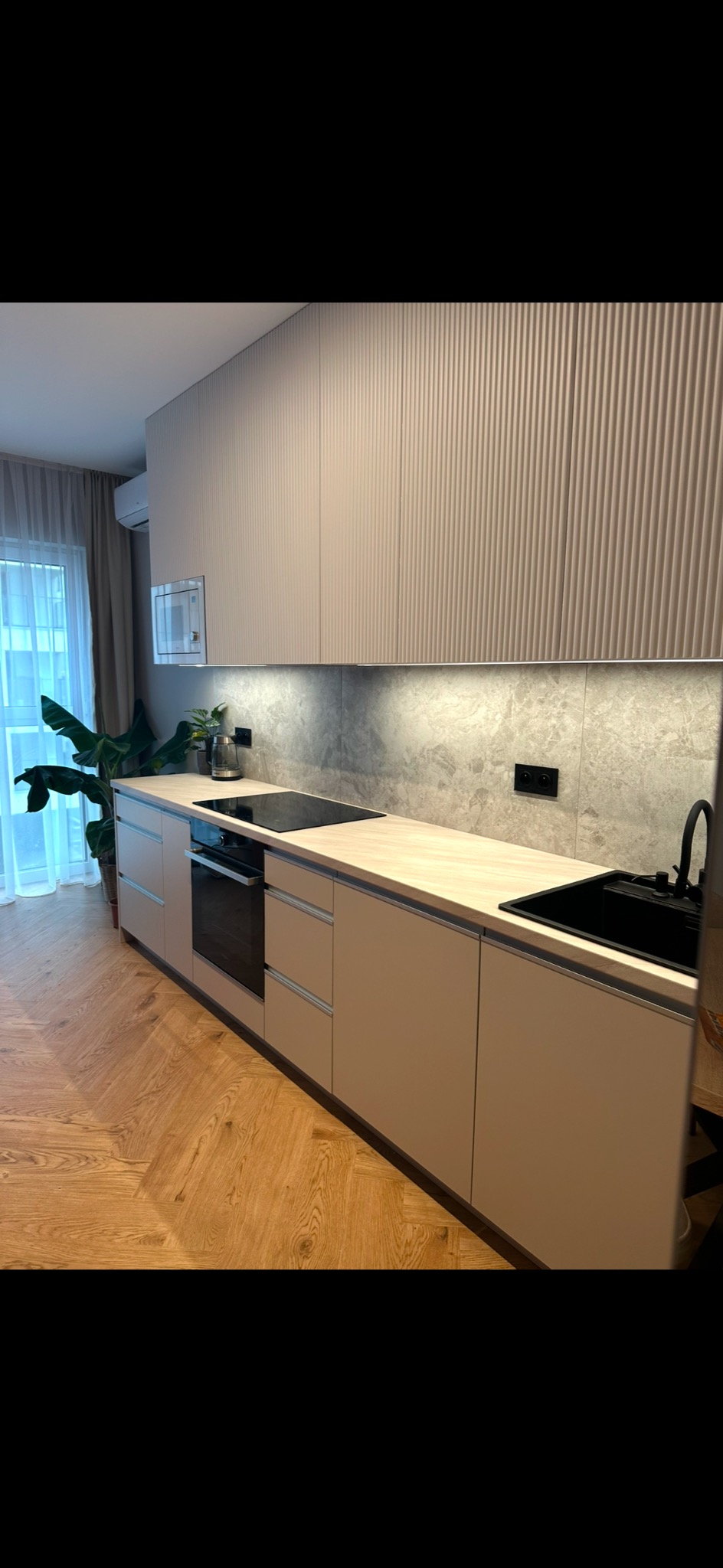 Beżowa kuchnia na wymiar z frezowanymi frontami, czarnym zlewem i płytą indukcyjną, oświetlona listwą LED, z parkietem w jodełkę na podłodze i rośliną doniczkową w tle.