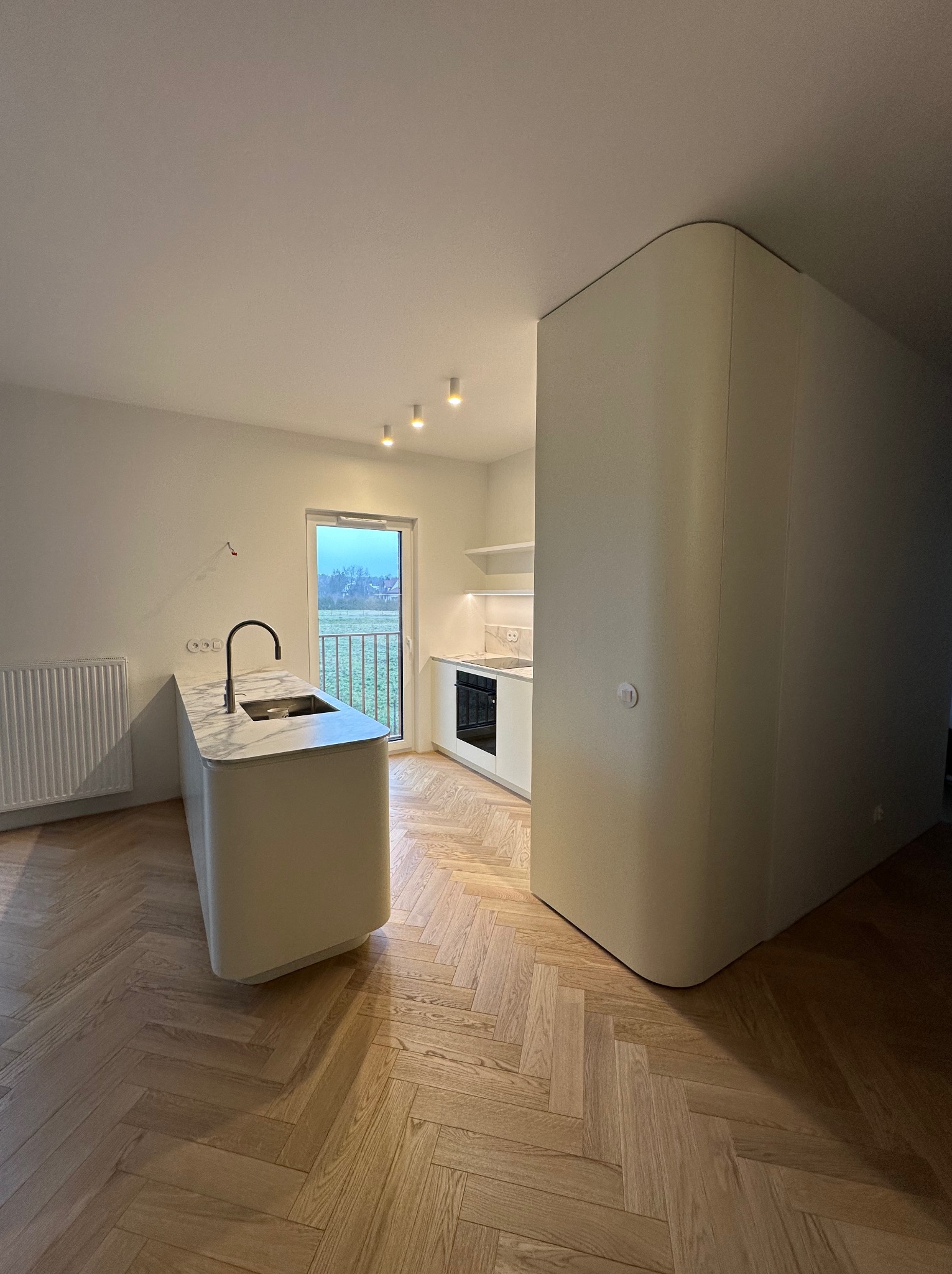 Minimalistyczna kuchnia z wyspą i marmurowym blatem, parkiet w jodełkę, widok na zewnątrz przez balkonowe drzwi.