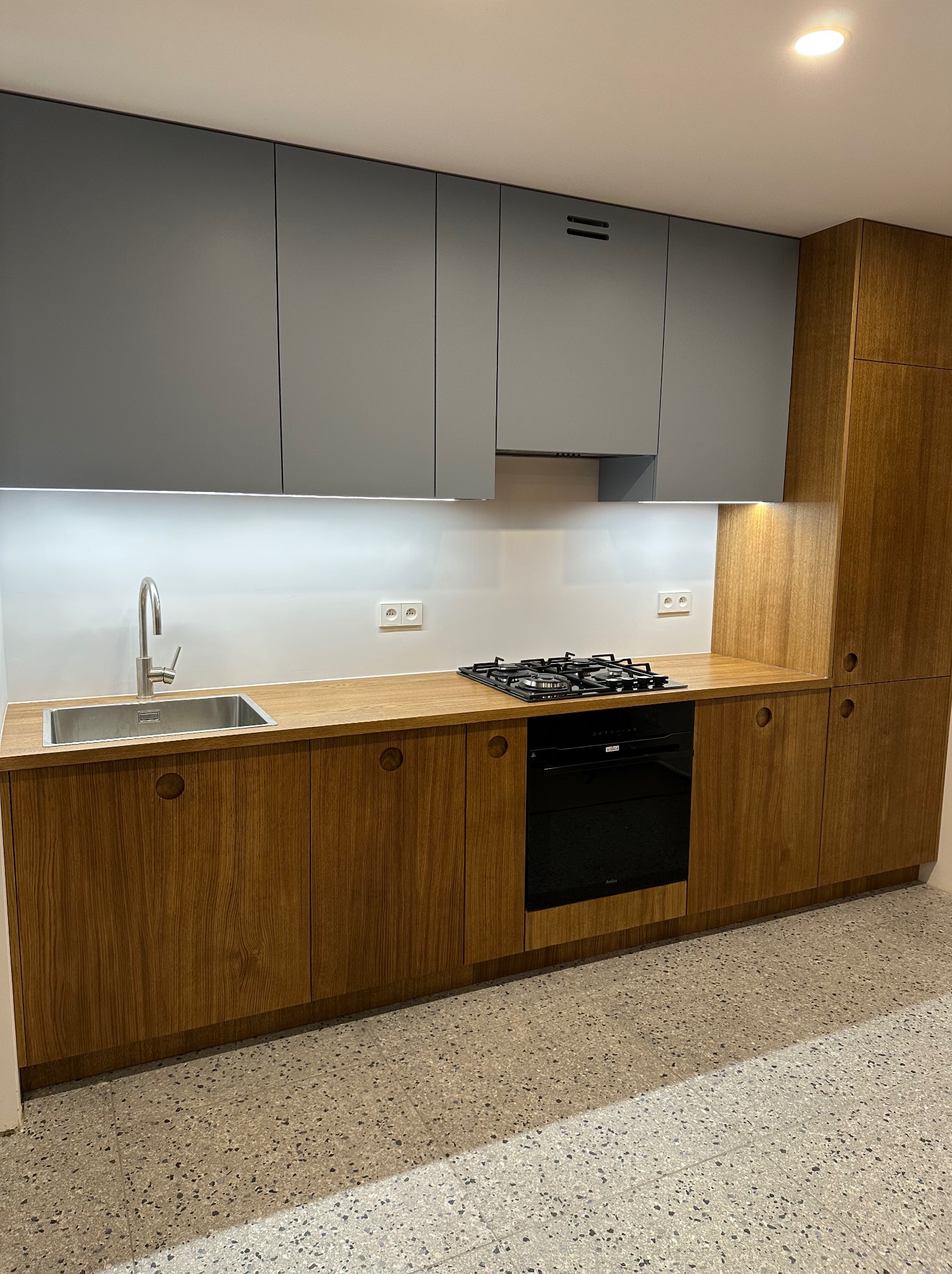 Nowoczesna kuchnia z szarymi górnymi szafkami i drewnopodobnymi dolnymi szafkami z okrągłymi uchwytami, zintegrowanym oświetleniem LED pod szafkami wiszącymi i płytą gazową.