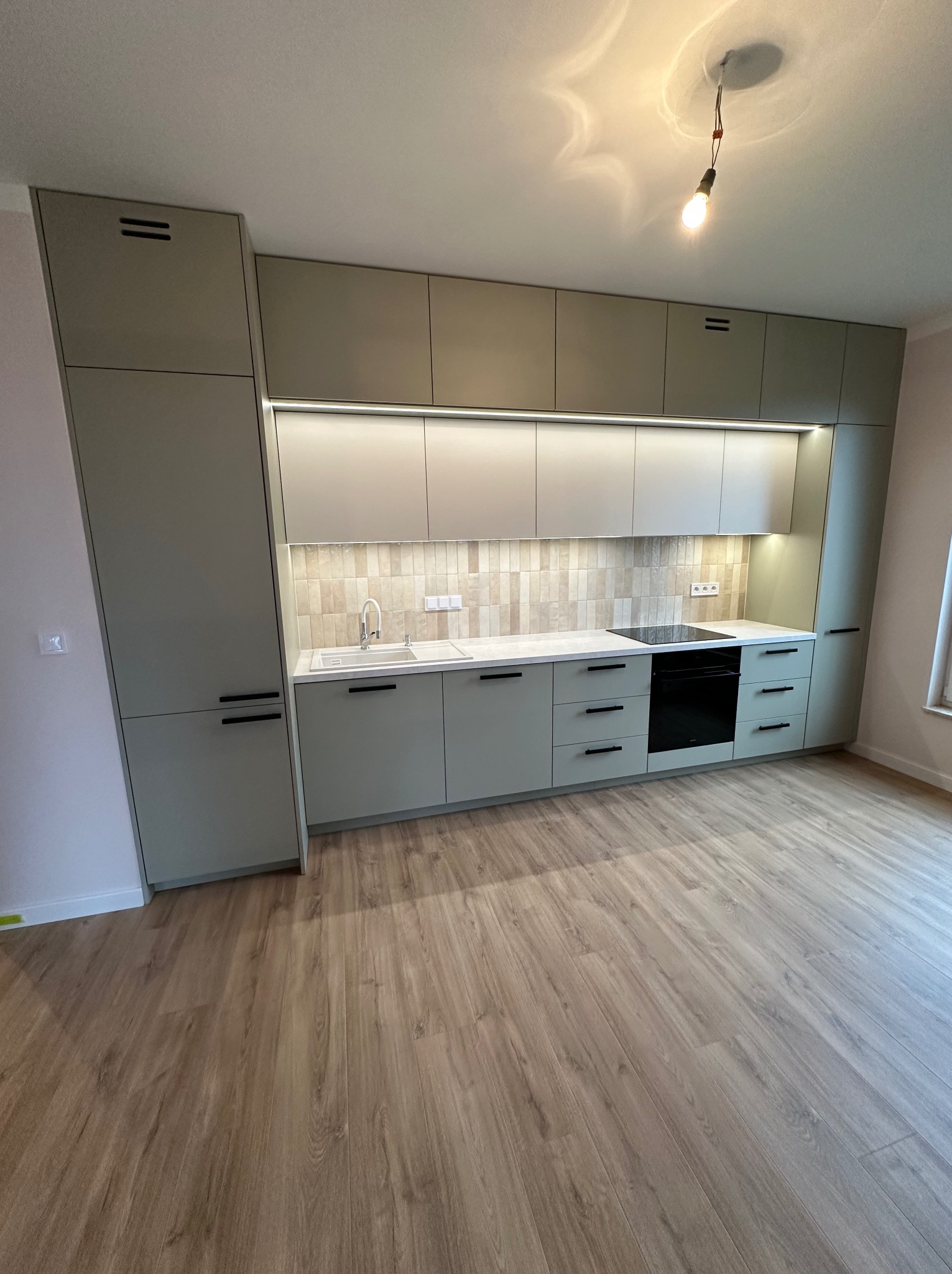 Jasna, nowoczesna kuchnia z zabudową w odcieniach szarości i beżu, z drewnianą podłogą i minimalistycznym oświetleniem podszafkowym.