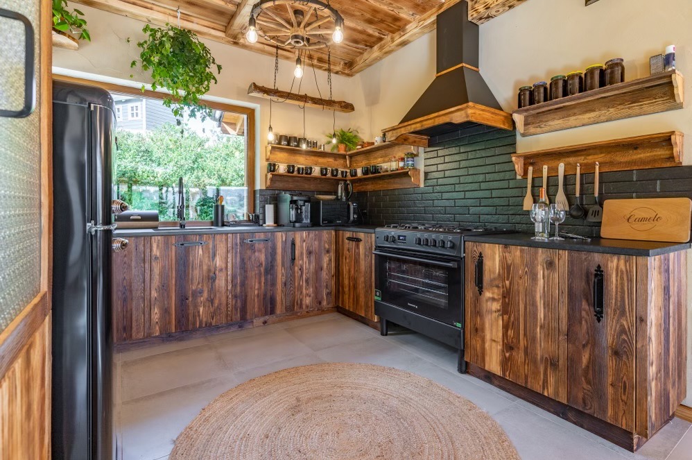 Rustykalna kuchnia z drewnianymi frontami szafek, czarnym sprzętem AGD i blatem, ceglaną ścianą nad blatem, drewnianym sufitem z dekoracyjnym kołem od wozu jako lampą, okrągłym jutowym dywanem...