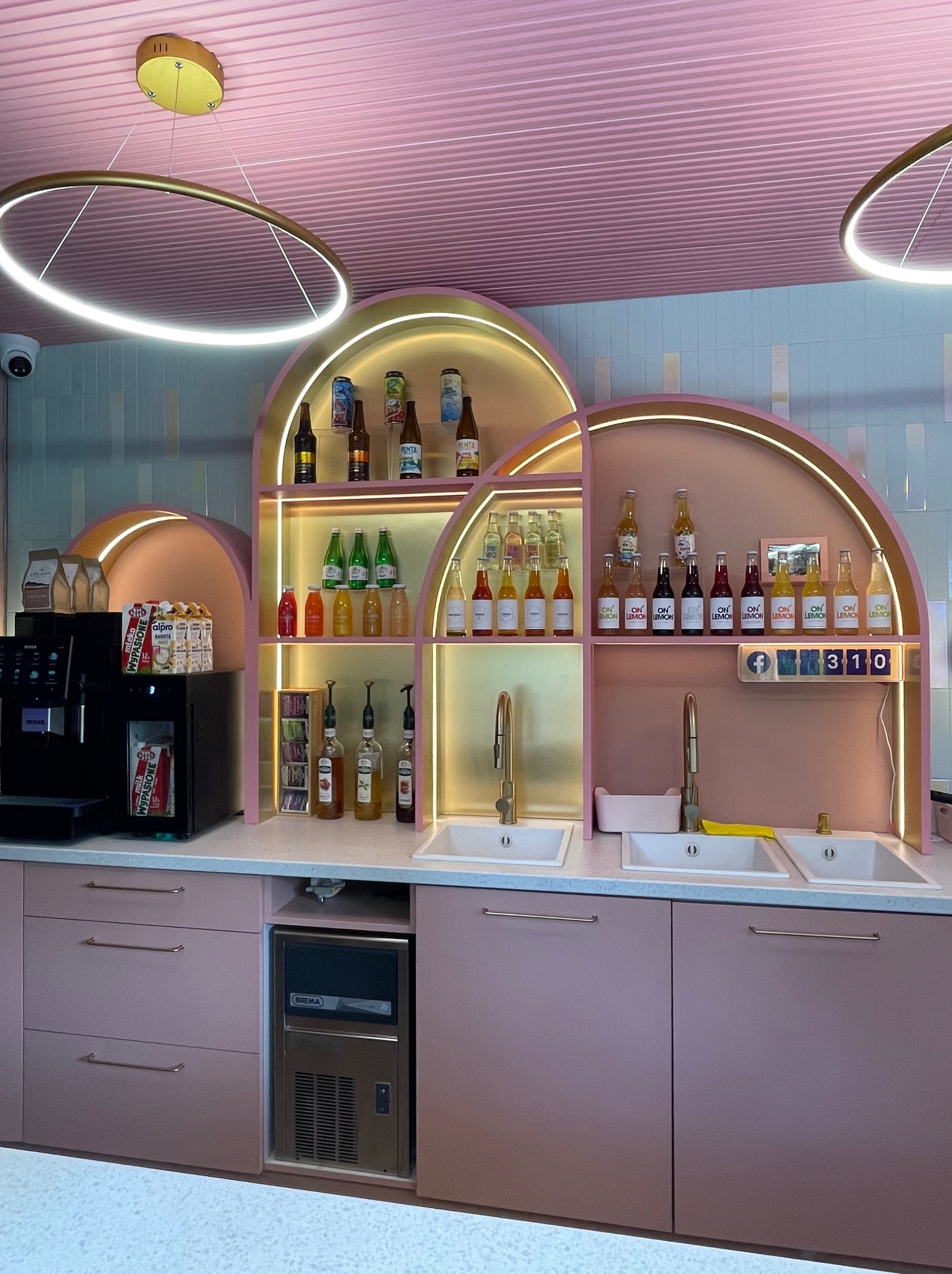 Różowa kuchnia komercyjna z półkami w kształcie łuków, wyposażona w ekspres do kawy, lodówkę do lodu, trzy zlewozmywaki z bateriami w kolorze złotym i butelki napojów gazowanych na półkach