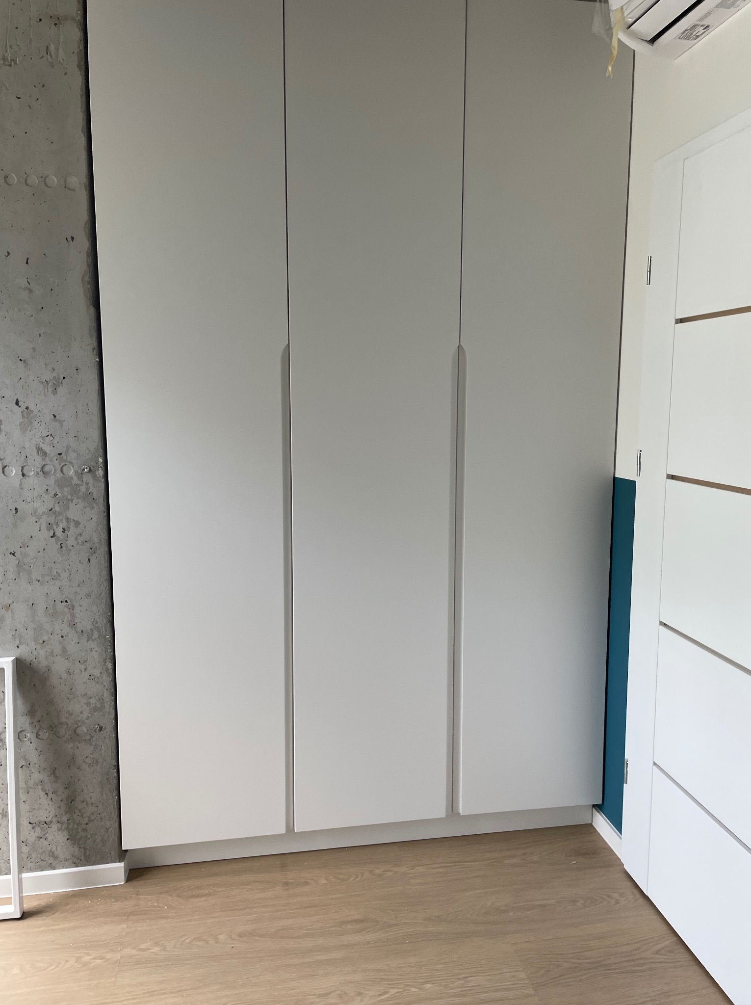 Trzydrzwiowa szafa wnękowa z białymi frontami, widoczna surowa ściana betonowa po lewej stronie i fragment drzwi po prawej stronie na tle drewnianej podłogi.