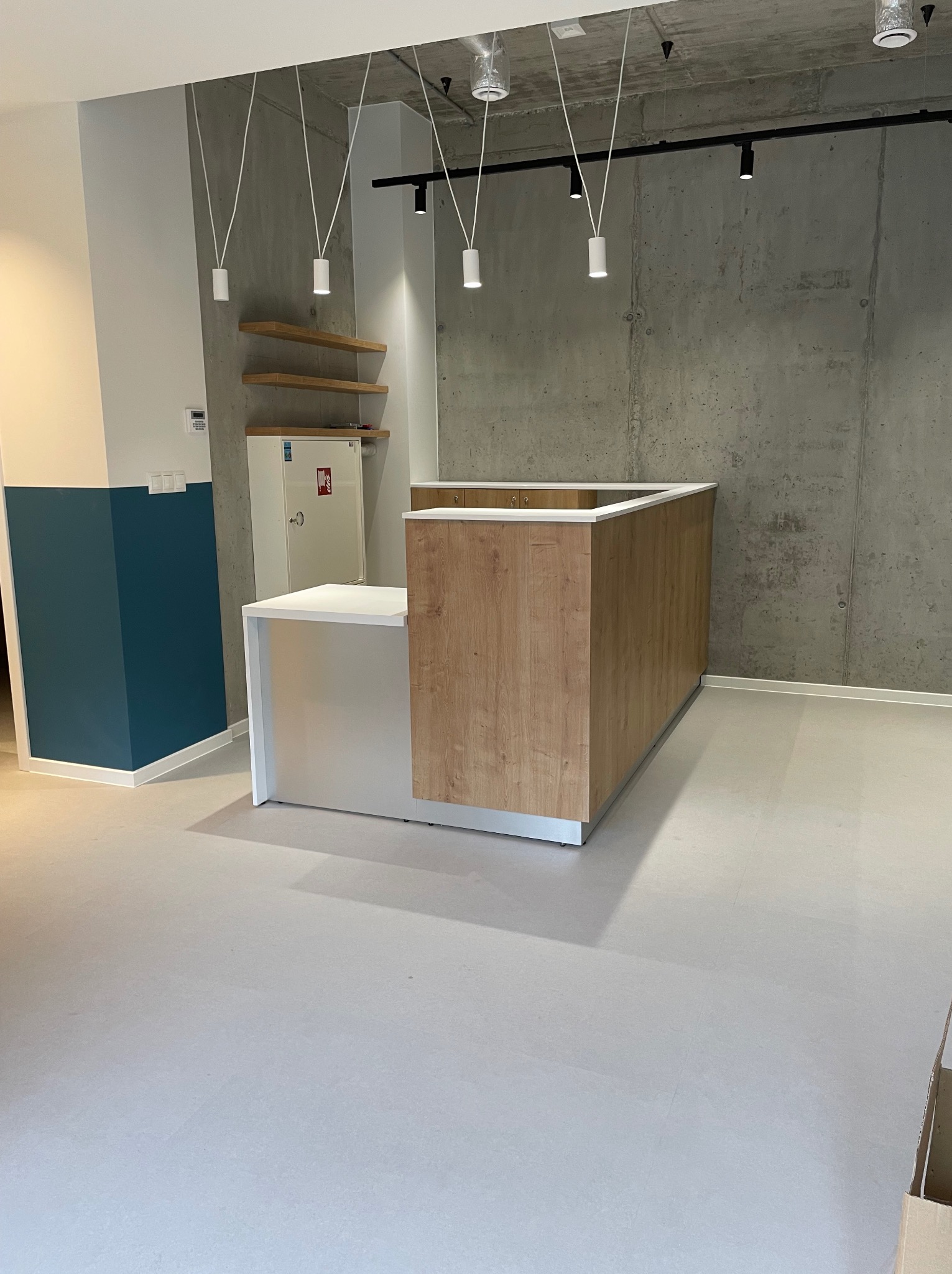Recepcja z blatem w kolorze białym i frontem w okleinie drewnopodobnej, widoczna na tle surowej, betonowej ściany i wiszących lamp