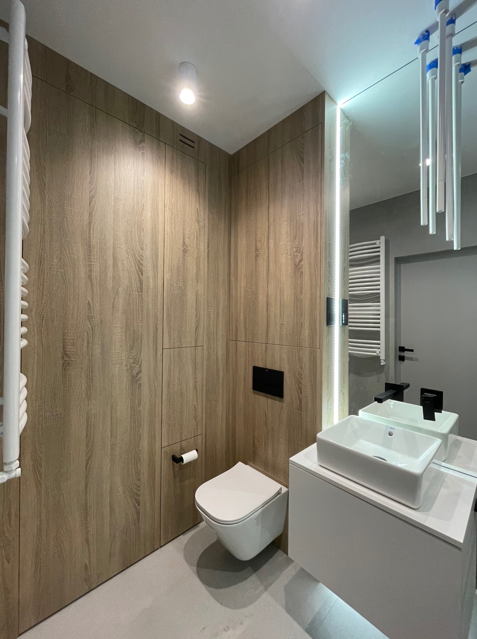 Nowoczesna łazienka z drewnianą zabudową meblową, podwieszaną toaletą i białą umywalką na szafce, oświetlenie LED przy lustrze.
