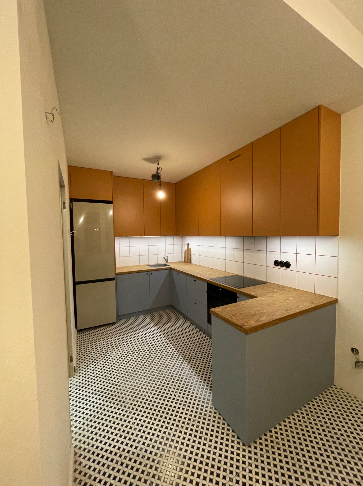 Ustawna kuchnia w stylu retro z brązowymi szafkami górnymi, szarymi dolnymi i blatem z jasnego drewna. Na podłodze wzorzysta, czarno-biała mozaika.