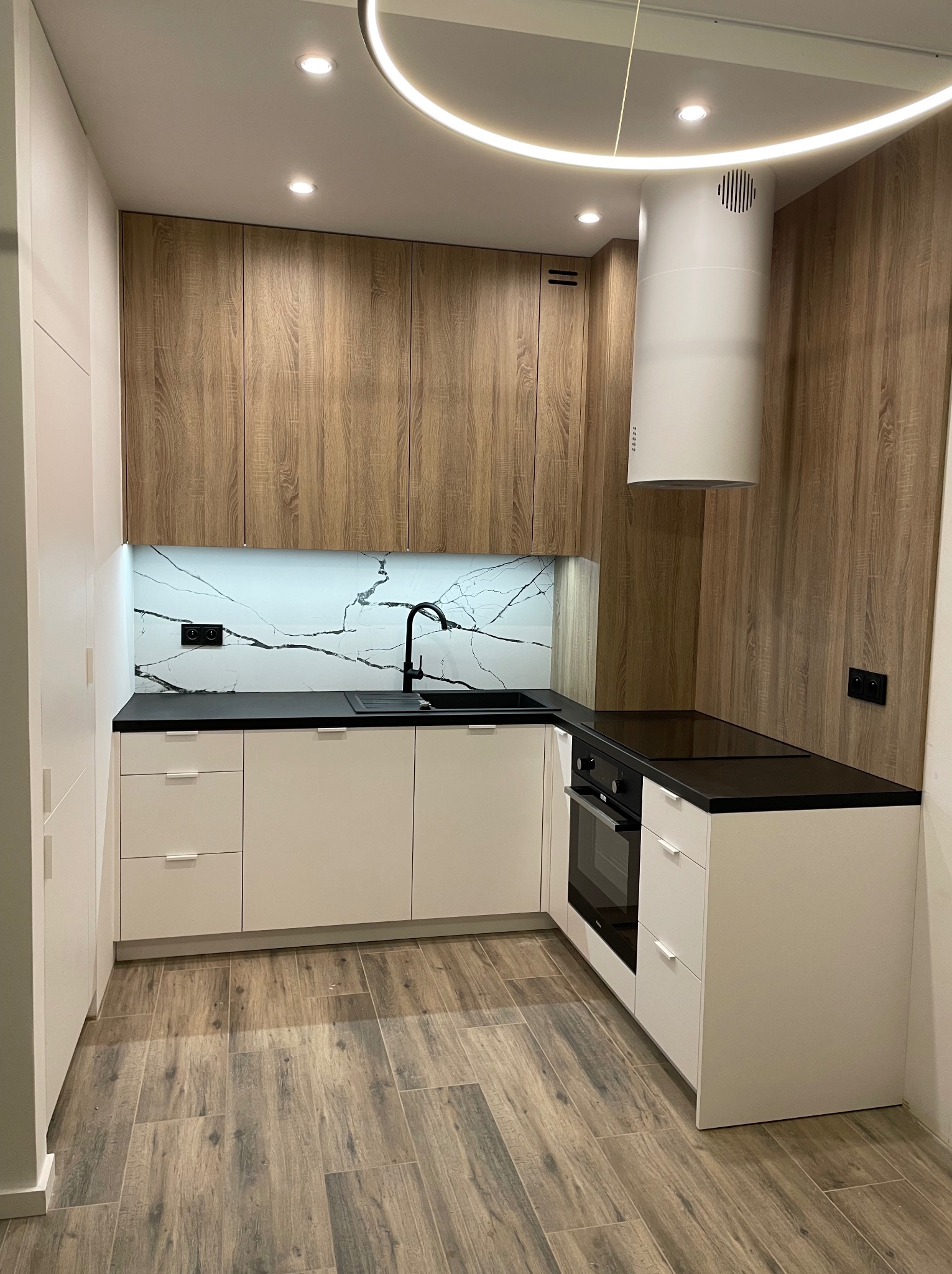 Nowoczesna kuchnia z białymi szafkami dolnymi, drewnopodobnymi szafkami górnymi, czarnym blatem i armaturą, oraz okapem cylindrycznym, oświetlona ledowym pierścieniem.