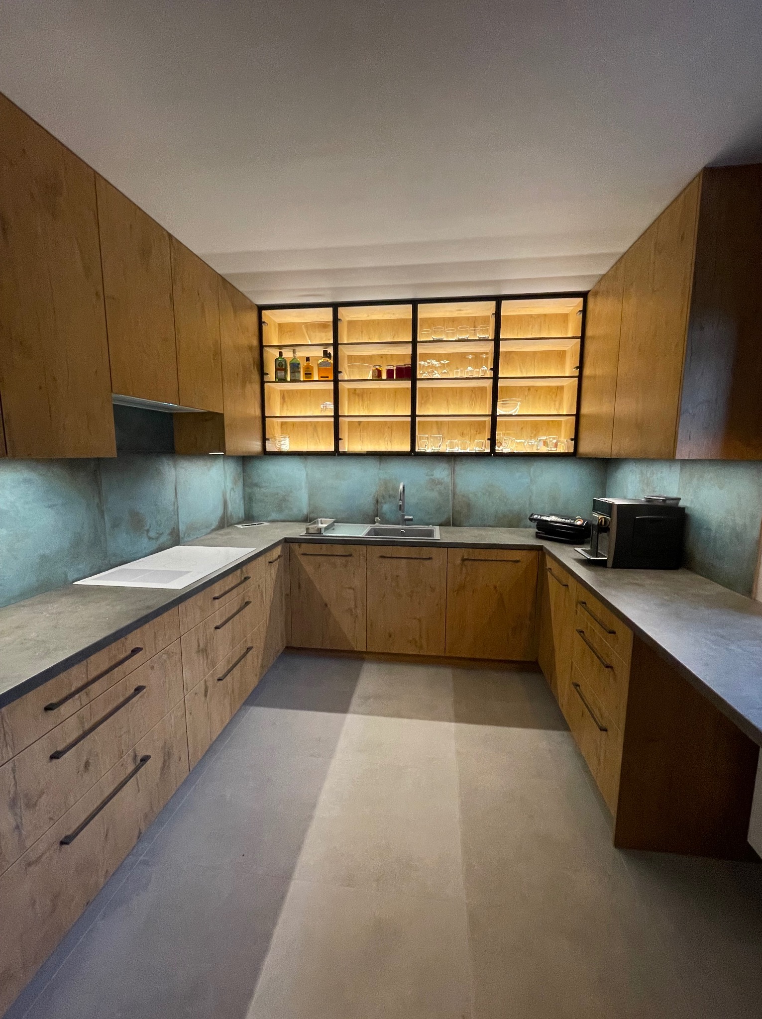 Nowoczesna kuchnia w kształcie litery U z drewnianymi frontami szafek, szarym blatem i podświetlaną półką z alkoholem i szkłem.