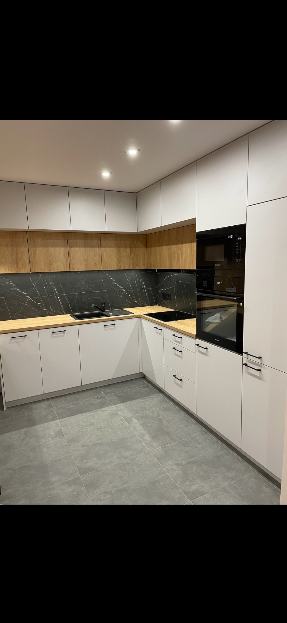 Nowoczesna kuchnia w kształcie litery L z białymi frontami, drewnianymi szafkami górnymi i blatem, czarnym marmurowym backsplashiem, czarnym zlewem i czarnymi uchwytami, oświetlona punktowymi...