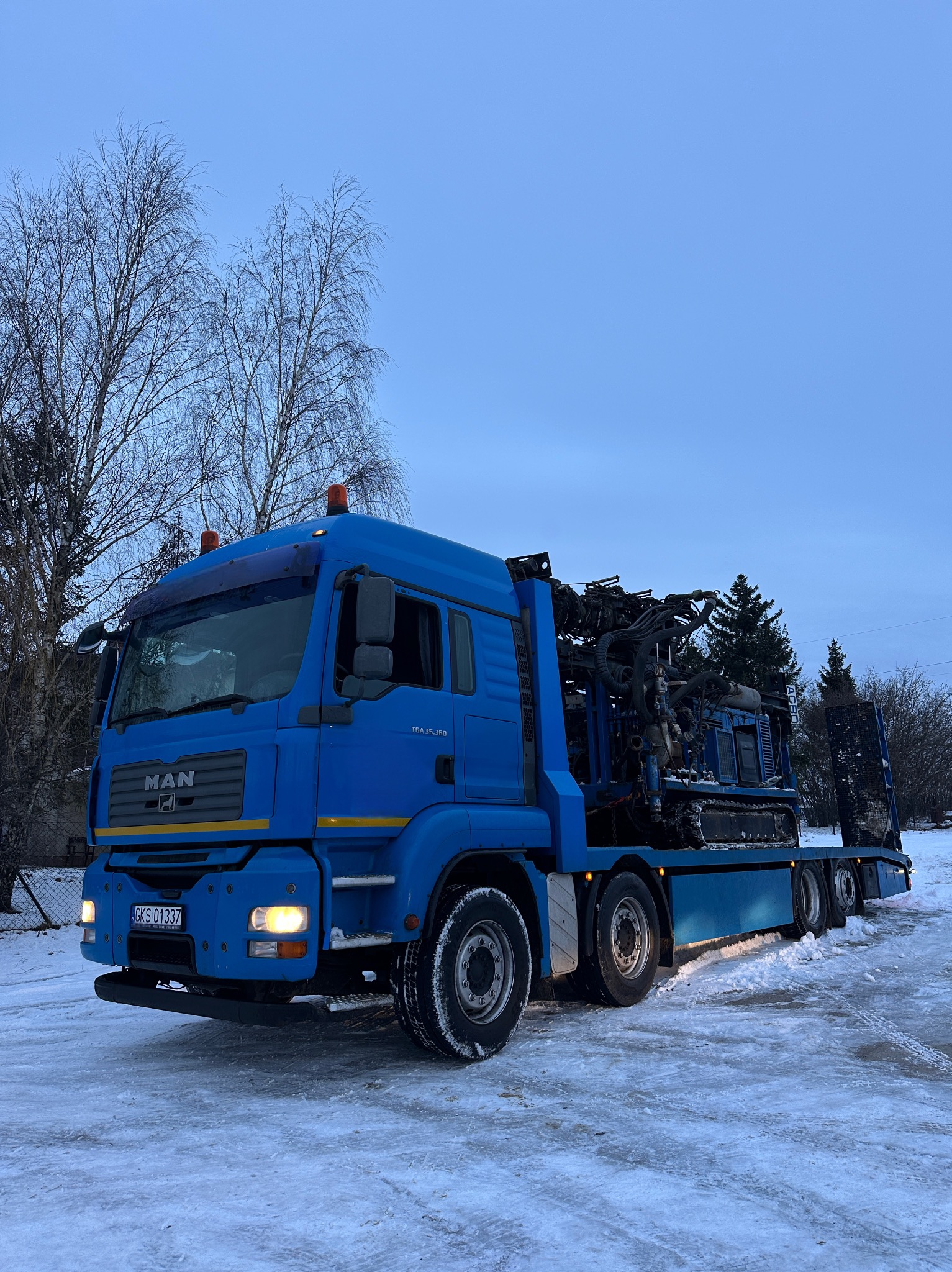 Niebieska ciężarówka MAN z wiertnicą na platformie, stoi na ośnieżonym terenie. W tle drzewa i błękitne niebo. Widoczne detale maszyny wiertniczej.