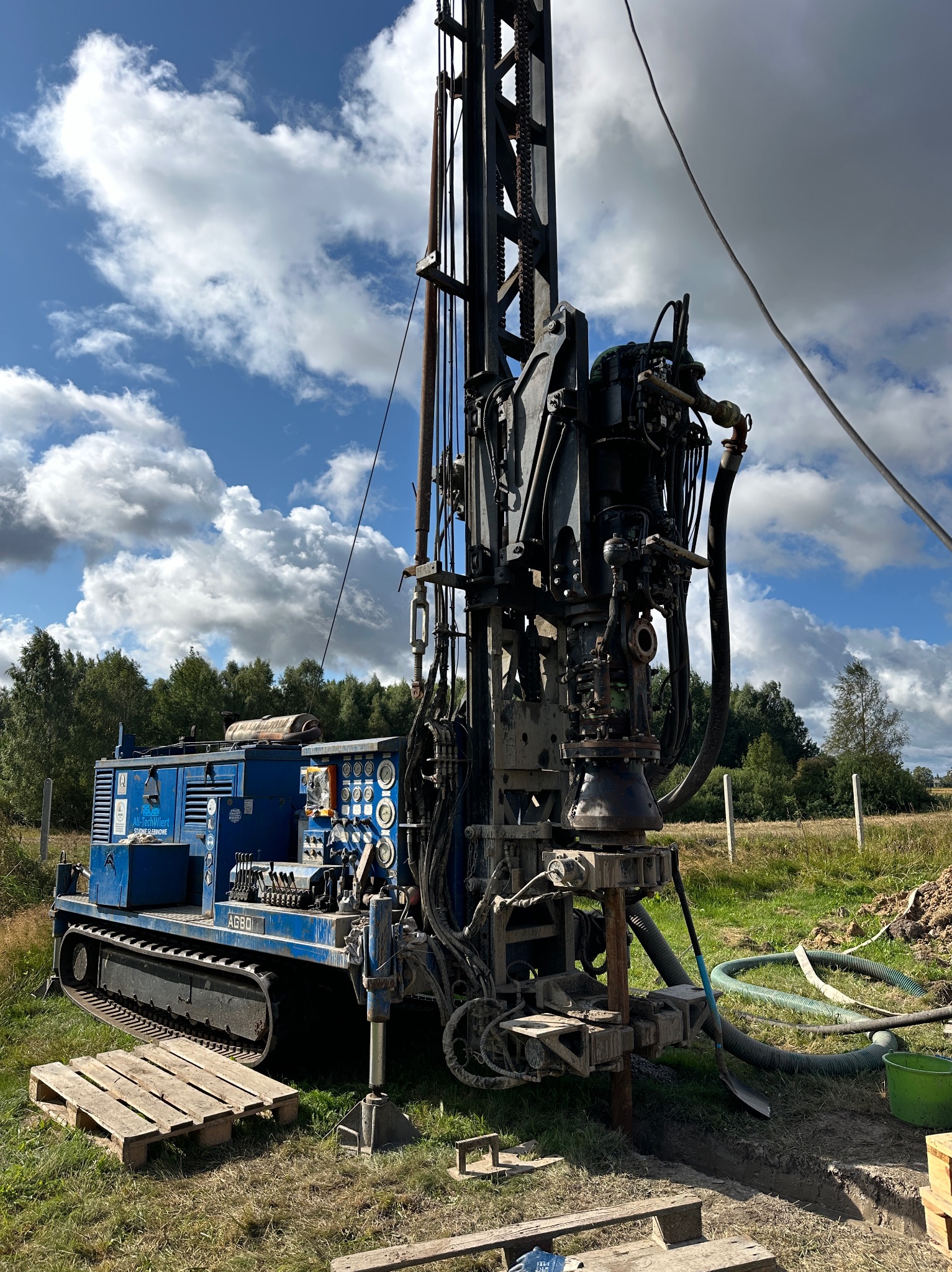 Wiertnica na gąsienicach, w trakcie pracy na zewnątrz, z widocznym wiertłem zagłębiającym się w ziemię. W tle błękitne niebo z chmurami i zieleń drzew.