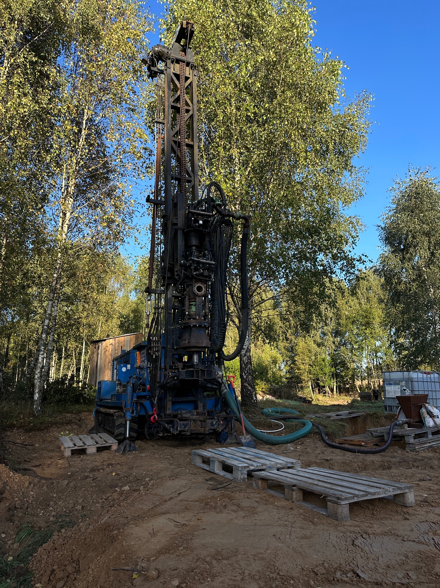 Wiertnica do studni głębinowych w Kościerzynie, na tle drzew i błękitnego nieba. Widoczne palety i zbiornik na wodę, teren przygotowany do wiercenia.