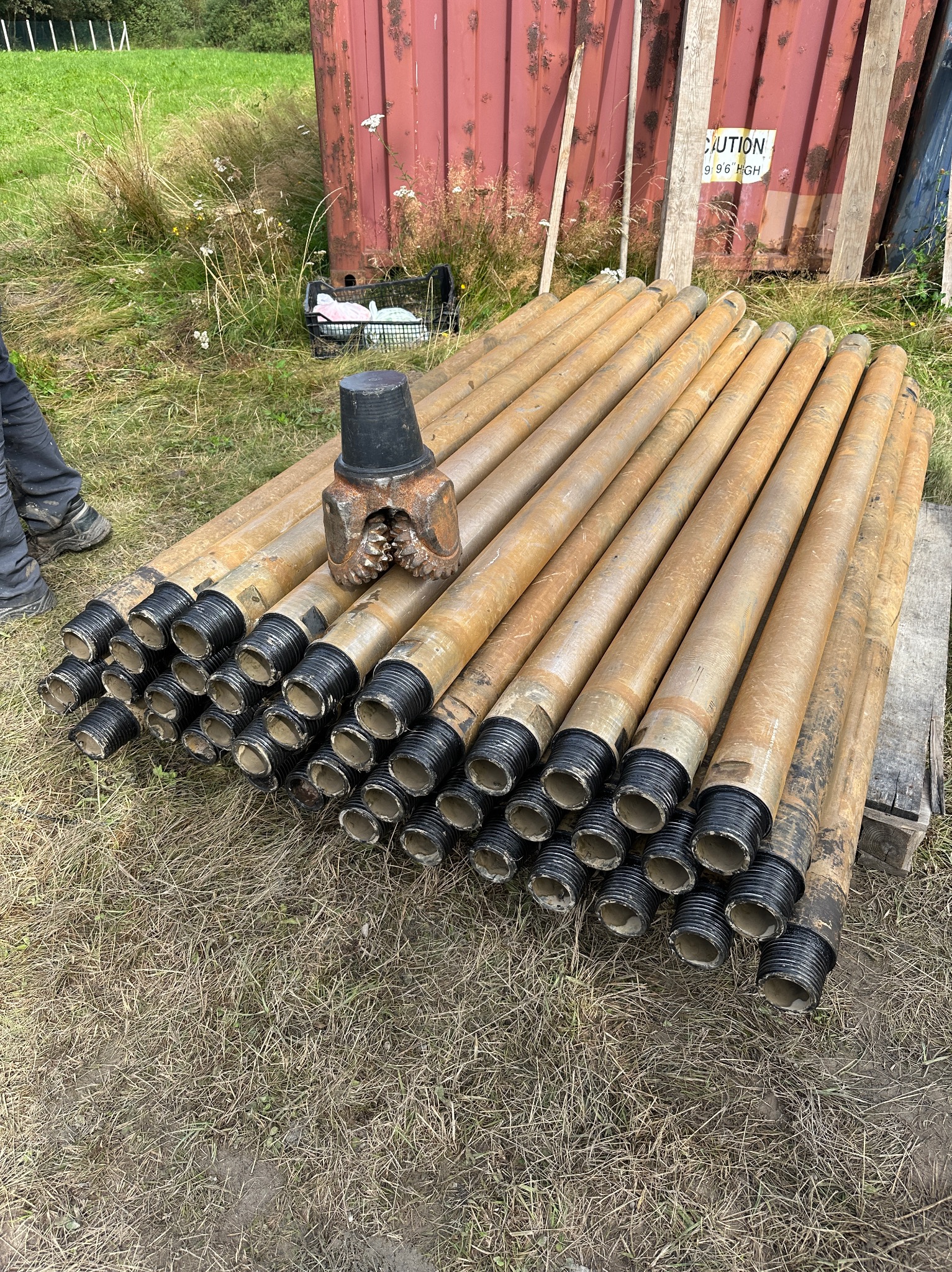 Stos rur wiertniczych z wiertłem na wierzchu, leżących na trawie obok czerwonego kontenera. Widoczne gwinty rur i zęby wiertła.