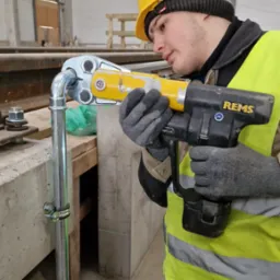 Hydraulik w żółtym kasku i kamizelce, zaciska złączkę na rurze za pomocą zaciskarki REMS, widoczny laserowy wskaźnik poziomu.