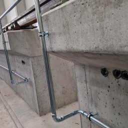 Włodek Hydraulik - Szare rury instalacji hydraulicznej zamocowane do betonowej ściany w obiekcie w Warszawie, widoczne otwory w ścianie i zabezpieczenie folią na podłodze.