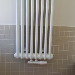 Włodek Hydraulik - Biały, żeberkowy grzejnik zamontowany na ścianie wyłożonej szarą mozaiką, z widocznym zaworem termostatycznym.