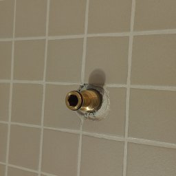 Włodek Hydraulik - Mosiężna końcówka instalacji hydraulicznej wystająca ze ściany z beżowych płytek, uszczelniona białą masą. Widoczny fragment instalacji z sześciokątnym otworem i gumową uszczelką.