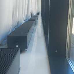 Włodek Hydraulik - Rząd czarnych, niskich grzejników kanałowych z kratką, umieszczonych wzdłuż białej ściany z zasłonami, widok z boku, oświetlenie dzienne.