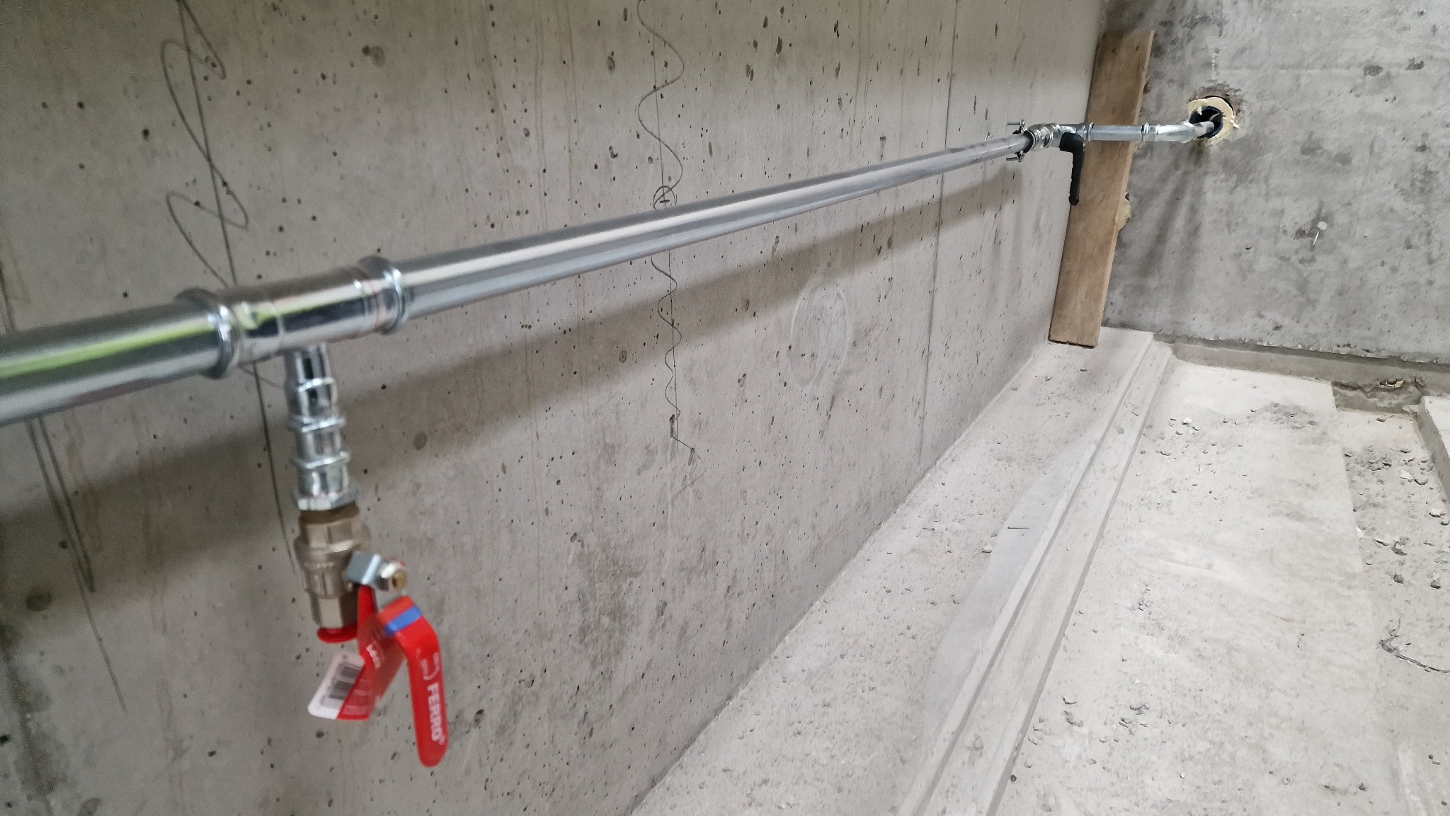 Szara ściana z betonu komórkowego z zamontowaną poziomą, chromowaną rurą instalacji wodnej z zaworem odcinającym z czerwoną dźwignią, widoczne ślady ołówka na ścianie i fragment posadzki.