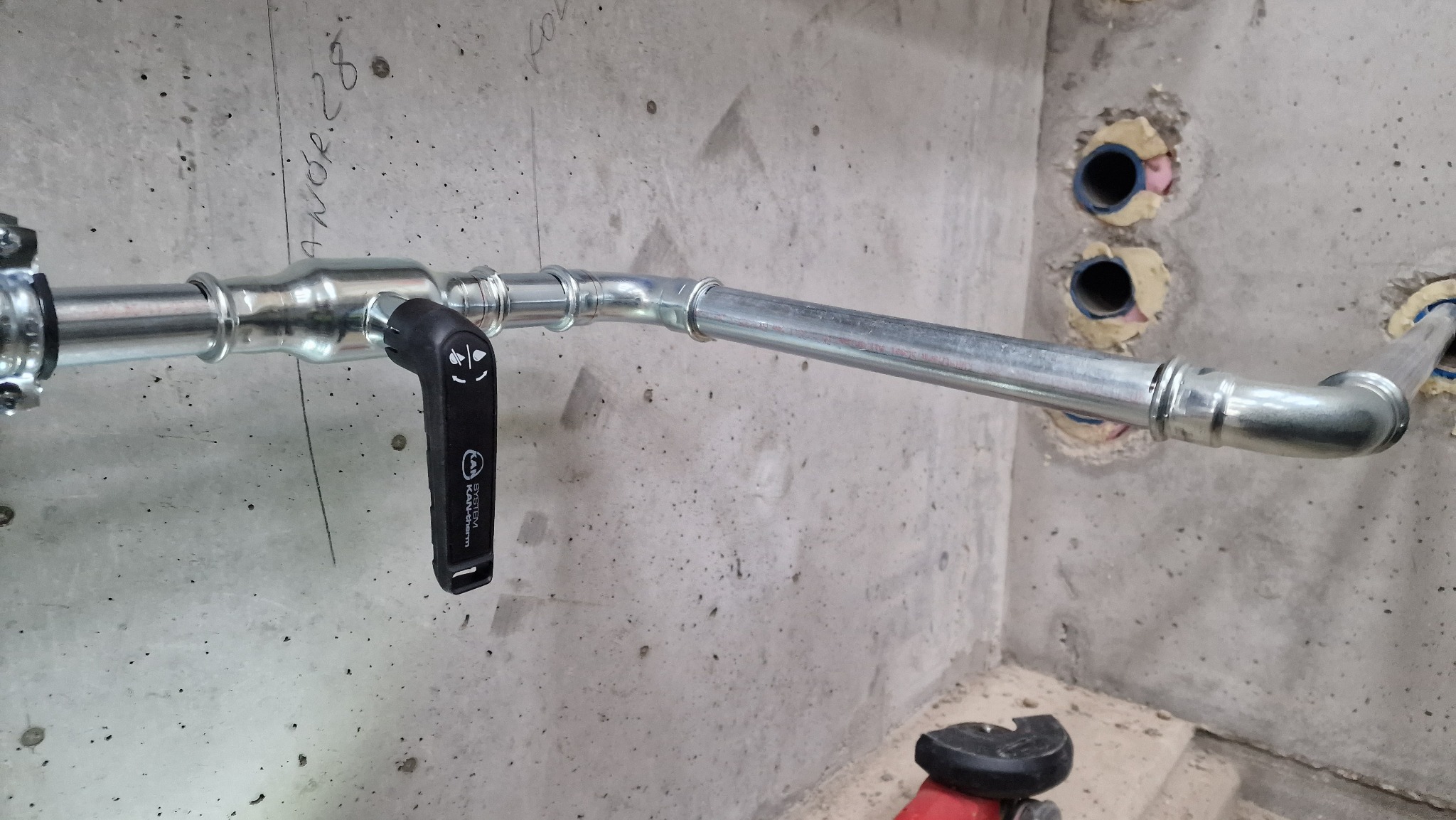 Szara, metalowa rura instalacji wodnej z czarnym zaworem kulowym, zamontowana na surowej ścianie betonowej z widocznymi otworami na inne rury, w tle fragment szlifierki kątowej.