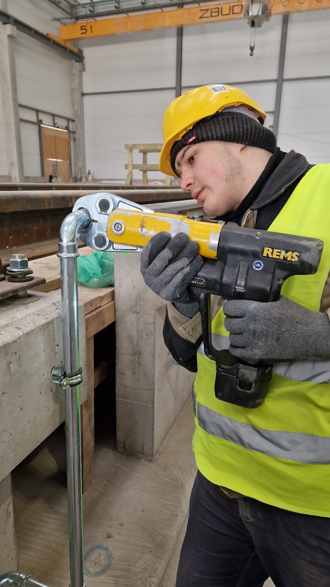 Hydraulik w żółtym kasku i kamizelce, zaciska złączkę na rurze za pomocą zaciskarki REMS, widoczny laserowy wskaźnik poziomu.