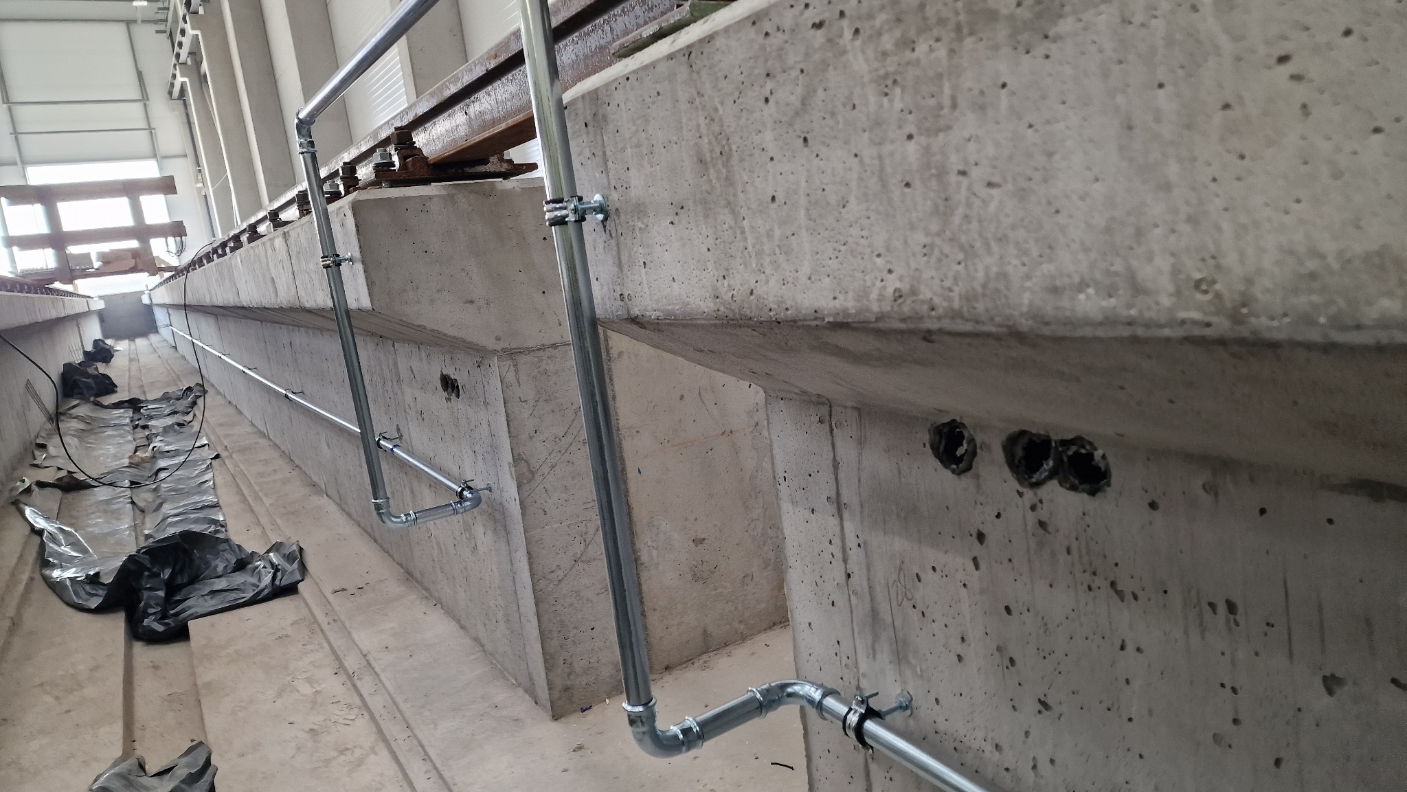 Szare rury instalacji hydraulicznej zamocowane do betonowej ściany w obiekcie w Warszawie, widoczne otwory w ścianie i zabezpieczenie folią na podłodze.