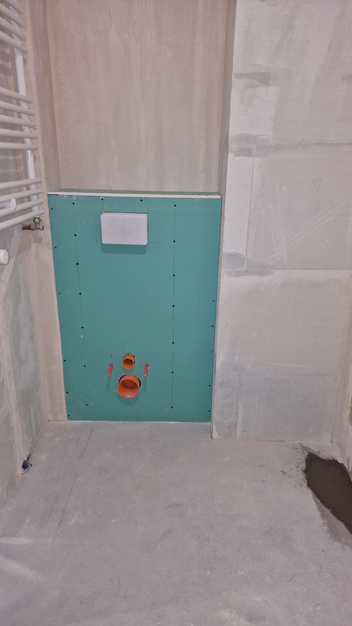 Ściana z płyty gipsowo-kartonowej w łazience w trakcie remontu, widoczne otwory na instalację podtynkową WC, rury kanalizacyjne w kolorze pomarańczowym, surowy beton na podłodze, grzejnik...