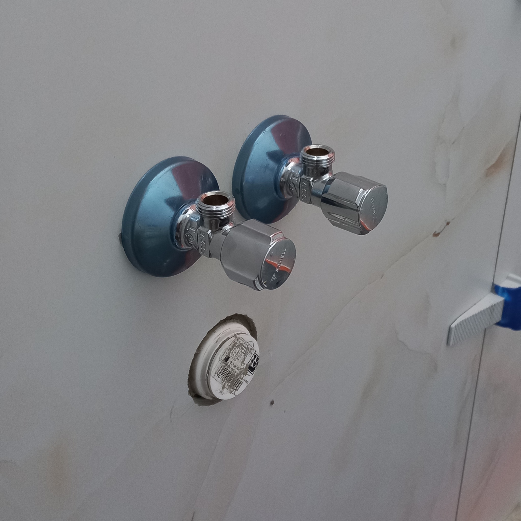 Dwa chromowane zawory kątowe Schell zamontowane na marmurowej ścianie, widoczna biała zaślepka z etykietą w otworze pod nimi, płytki wyrównywane niebieskim klipsem.