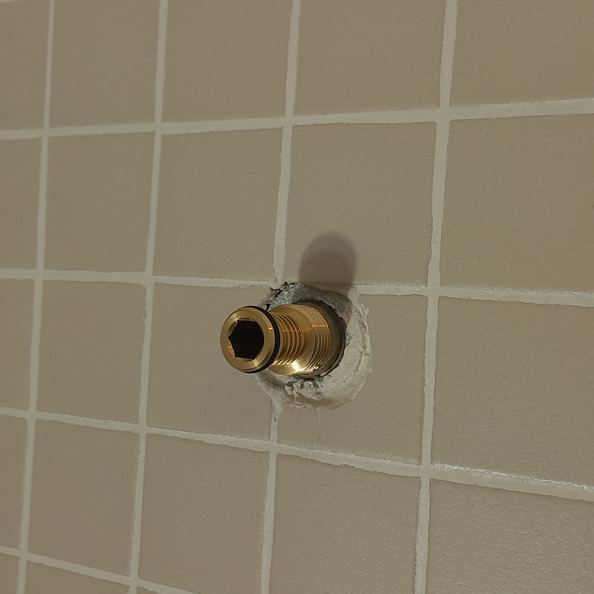 Mosiężna końcówka instalacji hydraulicznej wystająca ze ściany z beżowych płytek, uszczelniona białą masą. Widoczny fragment instalacji z sześciokątnym otworem i gumową uszczelką.