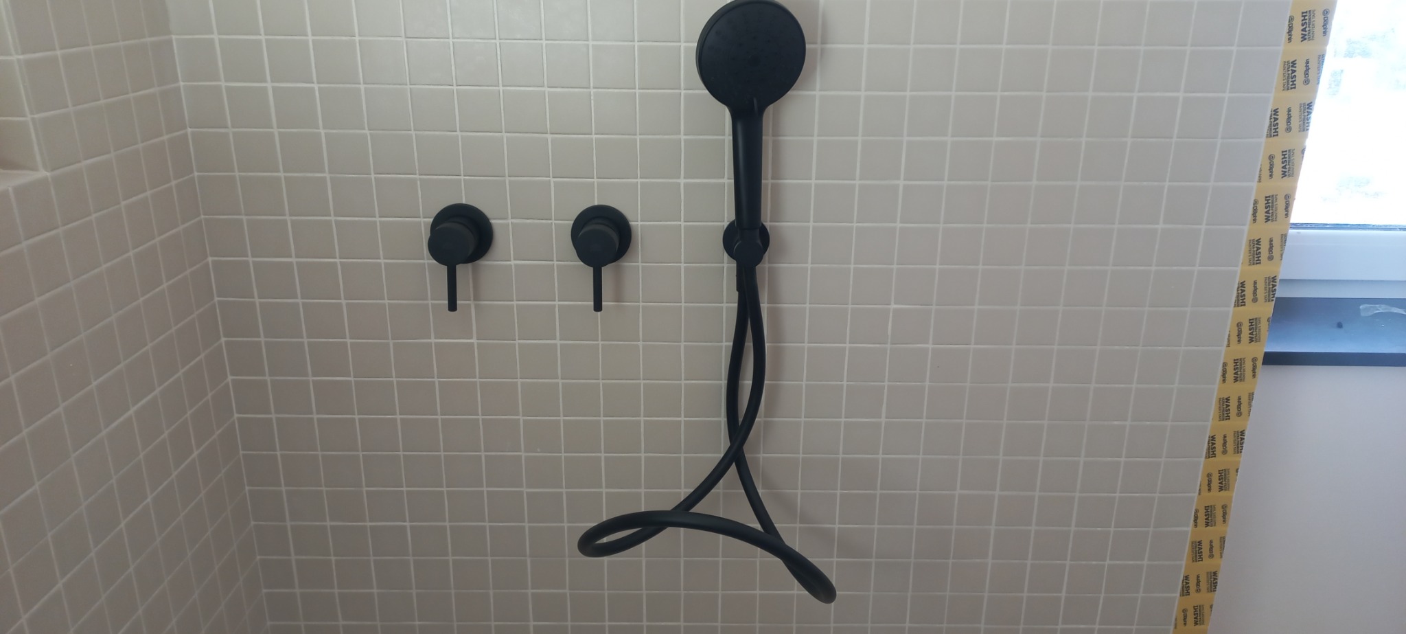 Czarna bateria prysznicowa podtynkowa z deszczownicą na ścianie wyłożonej jasnymi, kwadratowymi płytkami. Widoczne dwa pokrętła do regulacji wody. Przy oknie zabezpieczenie z taśmy malarskiej.