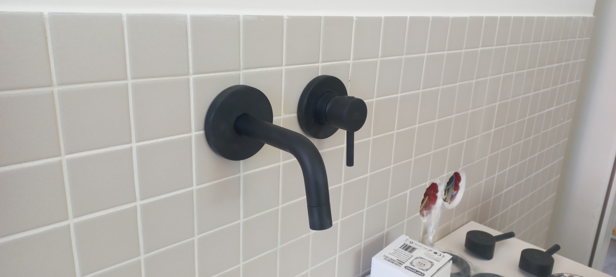 Bateria umywalkowa w kolorze czarnym, zamontowana na jasnych, kwadratowych płytkach. Widoczne podłączenia instalacji oraz akcesoria montażowe.