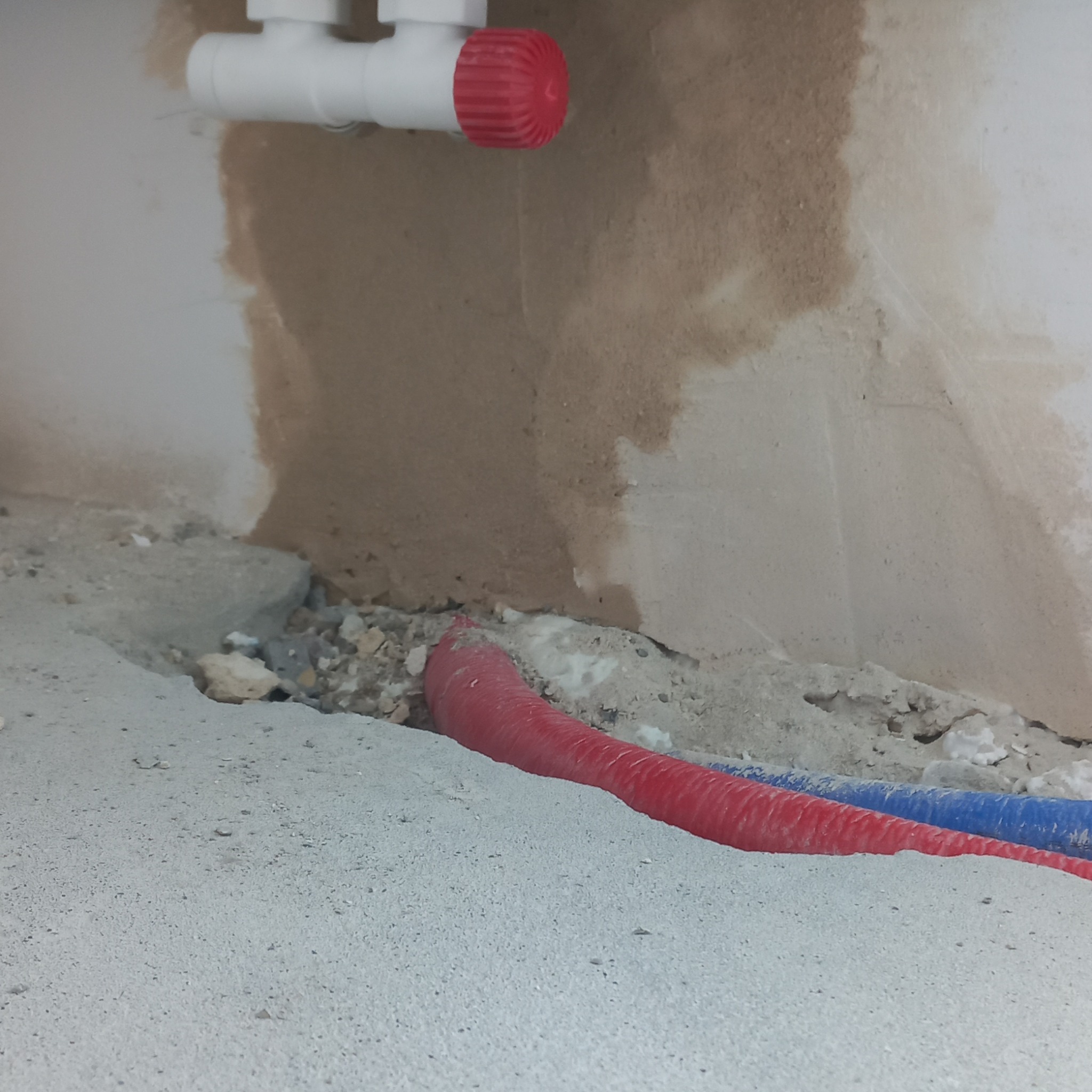 Fragment instalacji hydraulicznej w trakcie prac: czerwona i niebieska rura w wykopie w szarej posadzce, białe rury z czerwonym zaworem na ścianie z widocznymi śladami po kuciu.
