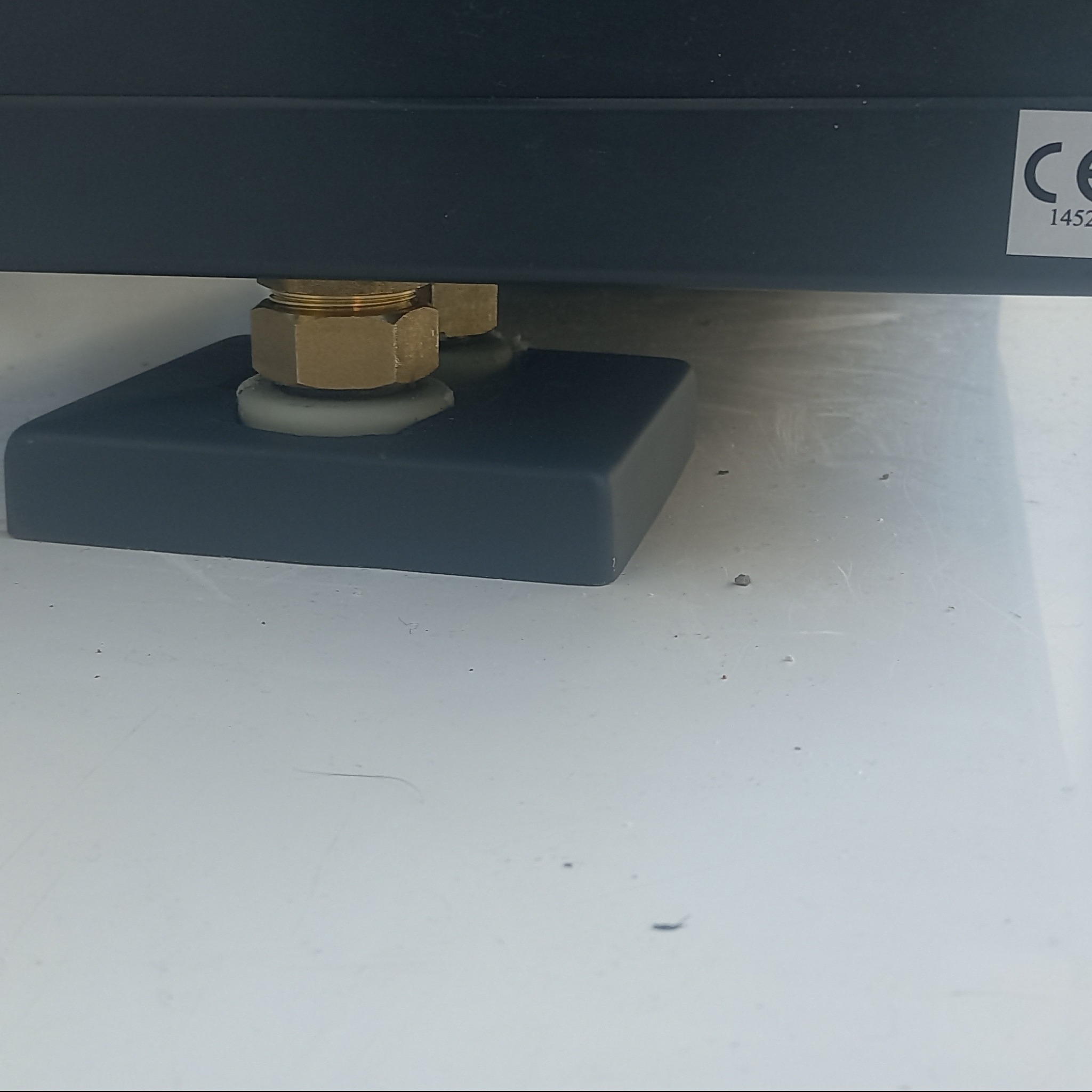 Szczegół mocowania pieca gazowego: mosiężne złączki, szara podkładka silikonowa i czarna podstawa na białej powierzchni. Widoczna naklejka z oznaczeniem CE.