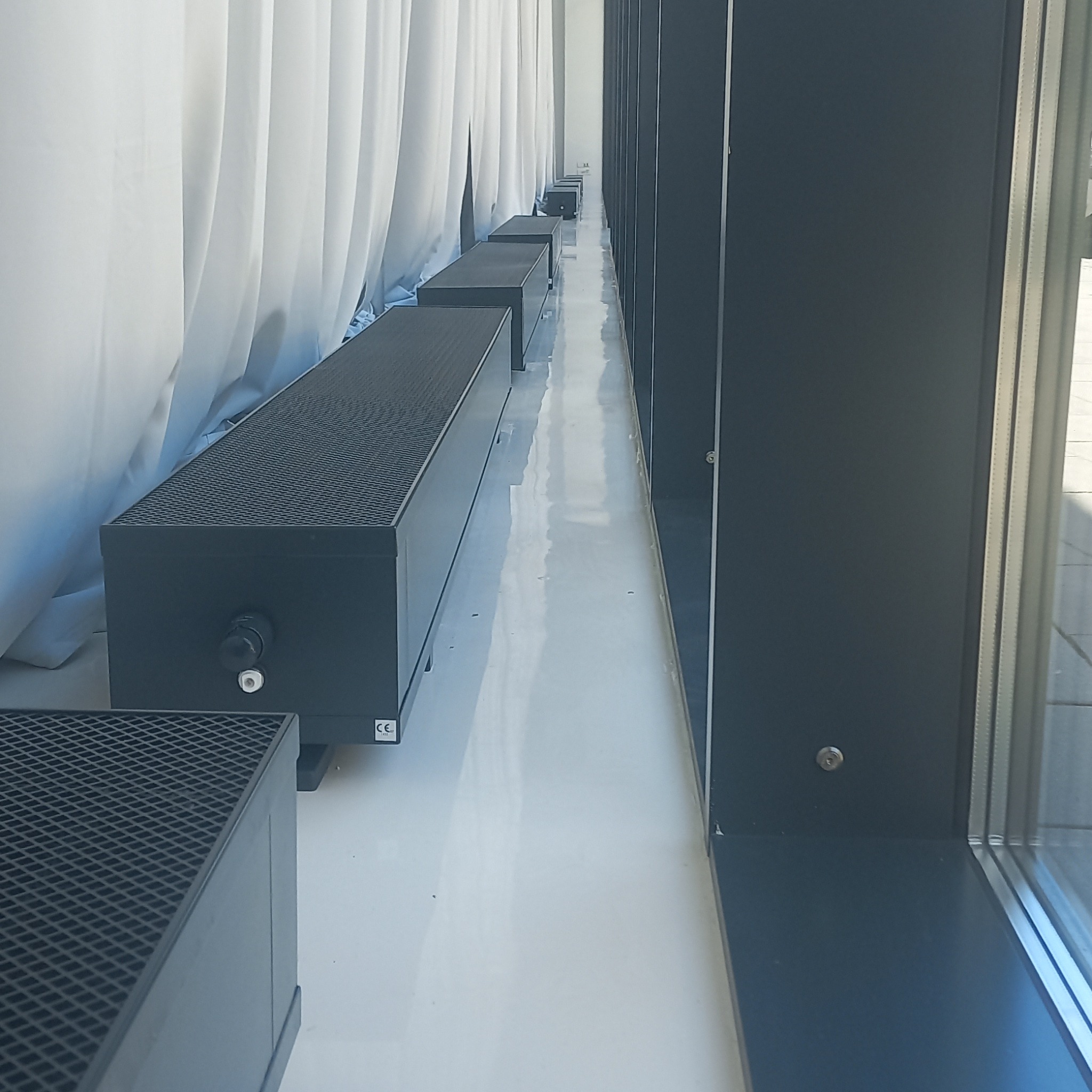 Rząd czarnych, niskich grzejników kanałowych z kratką, umieszczonych wzdłuż białej ściany z zasłonami, widok z boku, oświetlenie dzienne.