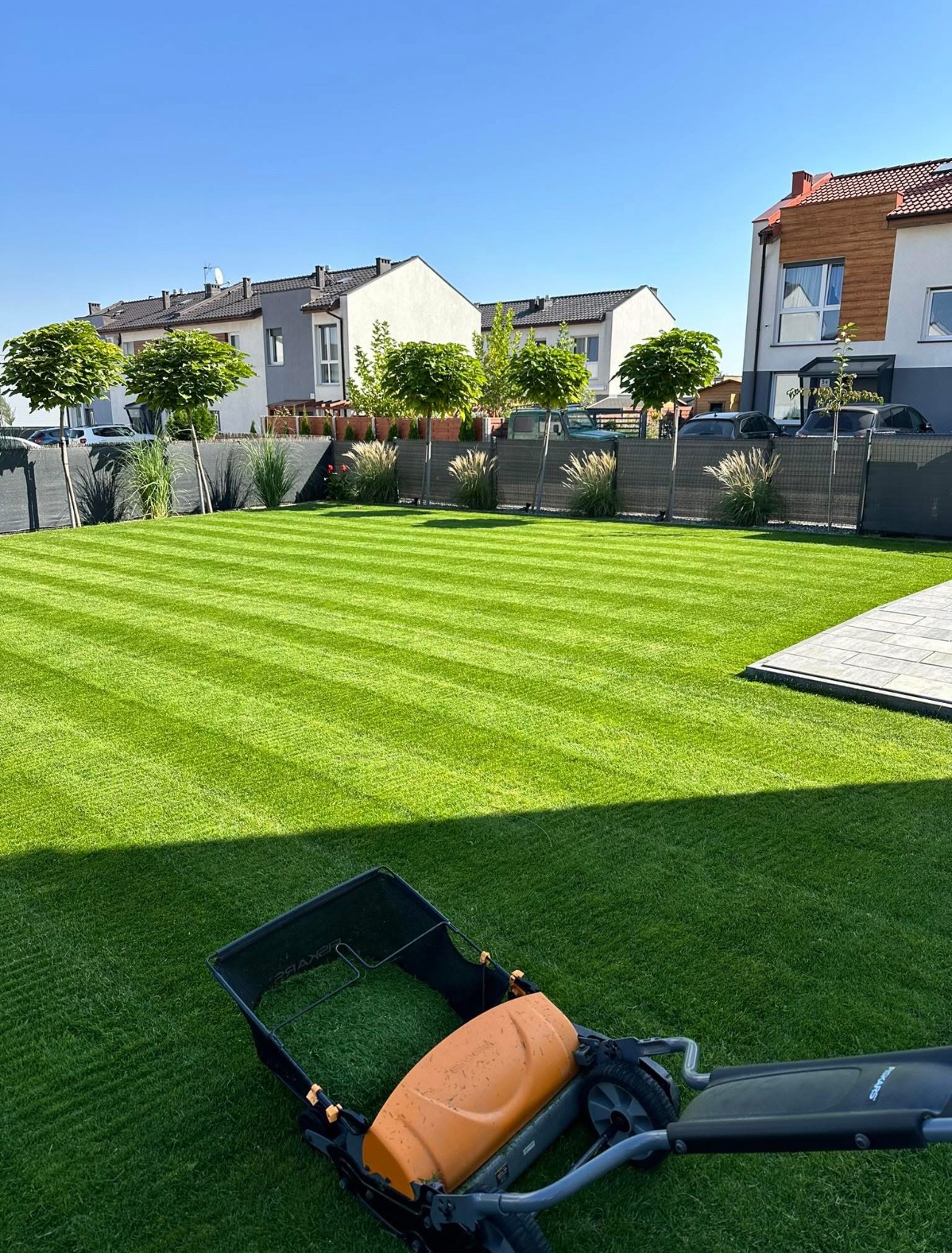 Perfekcyjnie skoszony trawnik z wyraźnymi pasami, z widoczną kosiarką Fiskars na pierwszym planie, w tle zadbane domy jednorodzinne i ozdobne drzewa.