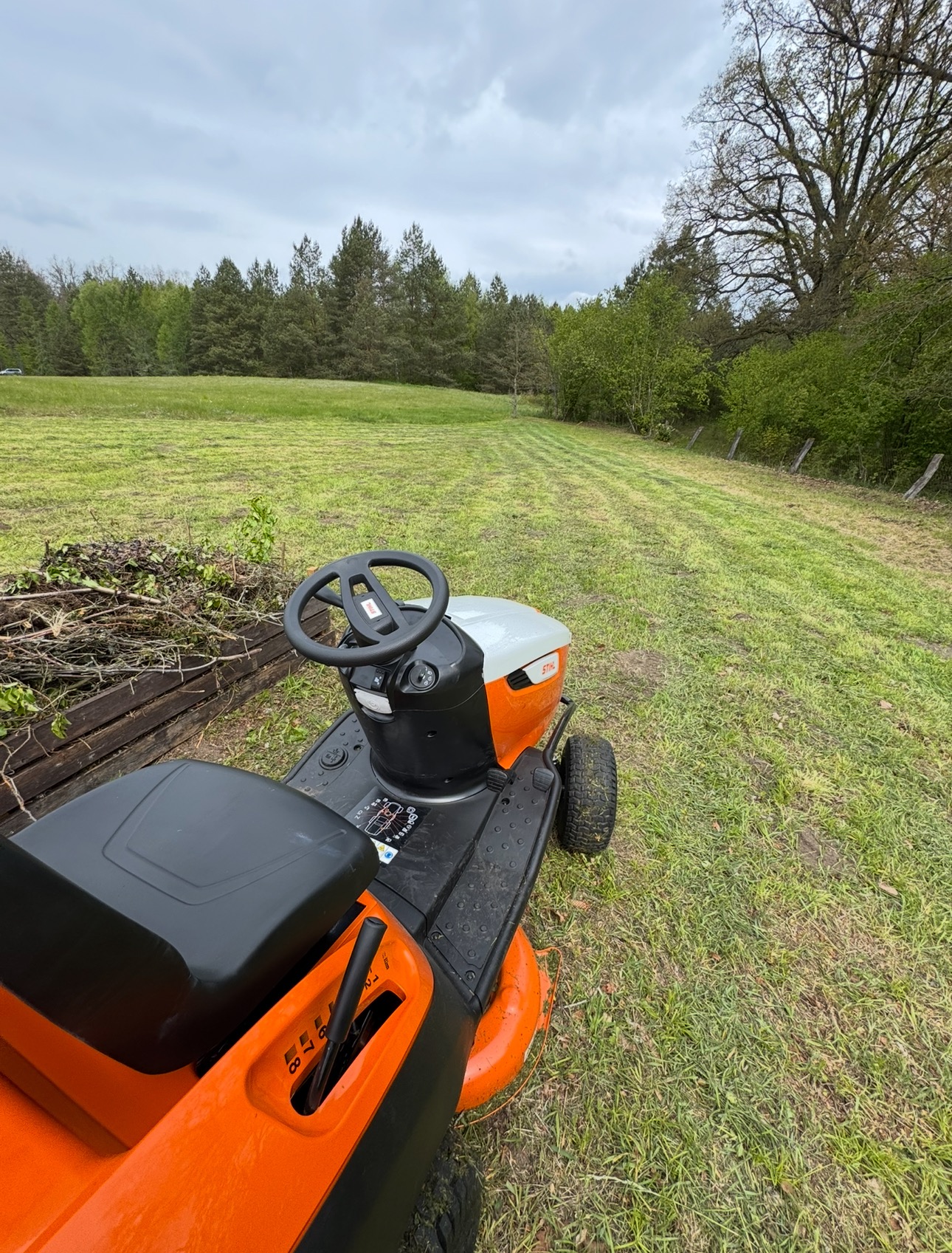 Traktorek do koszenia trawy Stihl na tle częściowo skoszonego trawnika w Jordanowie. Widoczne pasy skoszonej trawy i sterta gałęzi.