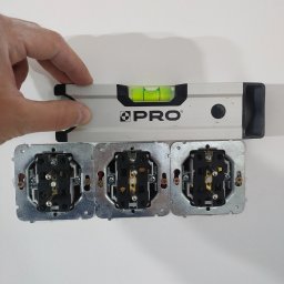Przemysław Łuczka - Poziomica PRO przykładana do potrójnego gniazdka elektrycznego w trakcie montażu, widoczne przewody i elementy mocujące.